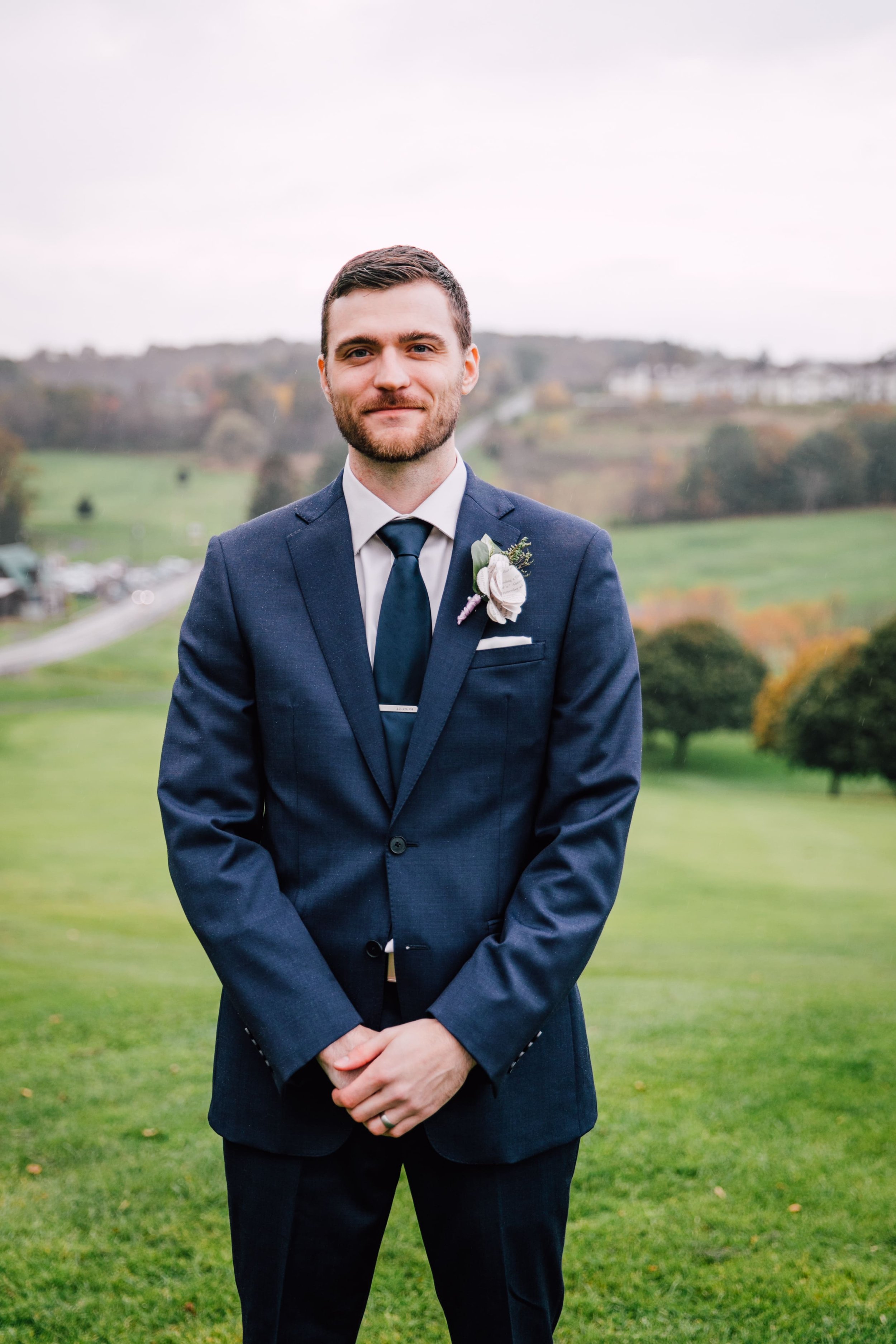 the groom smiles as he stands in front of an open field with fall trees behind him during their fall wedding portraits 