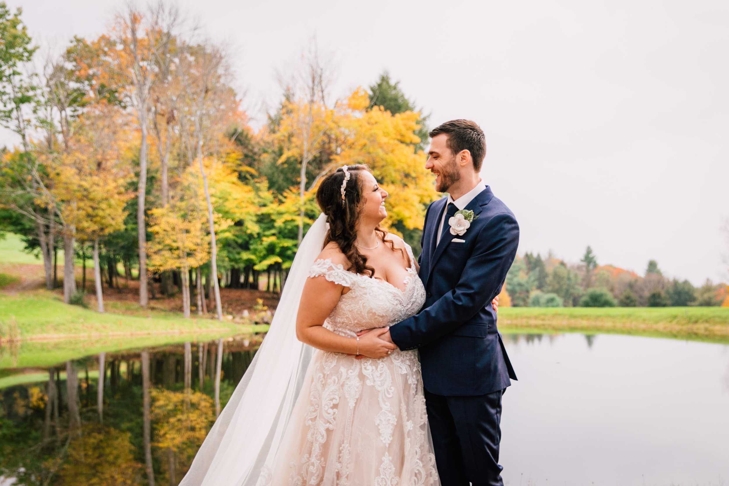  the bride and groom smile genuinely at each other as they stand in front of a small pond and fall foliage during their fall wedding photos 
