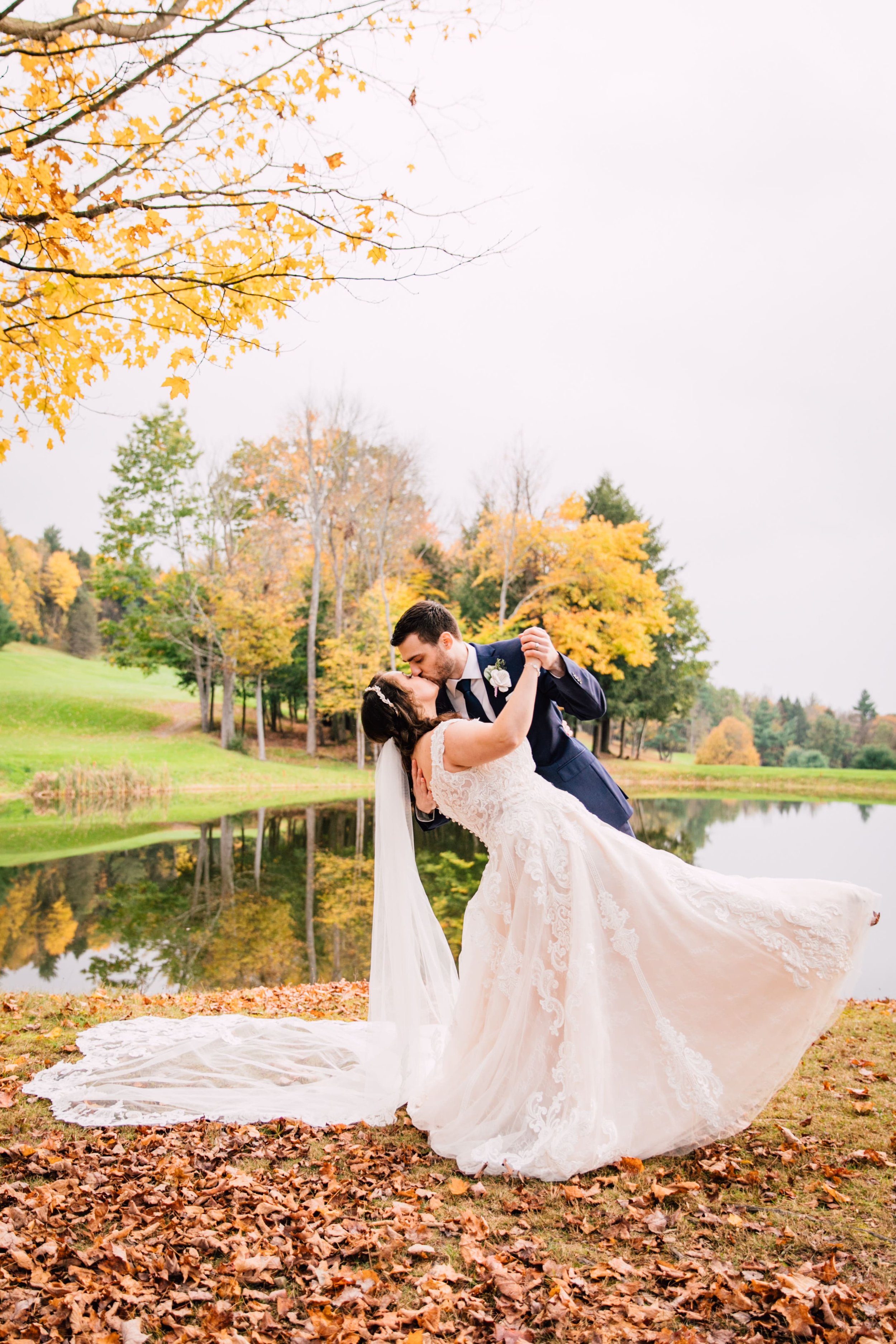  the groom dips the bride as they dance in front of a pond and kiss while taking fall wedding photos 