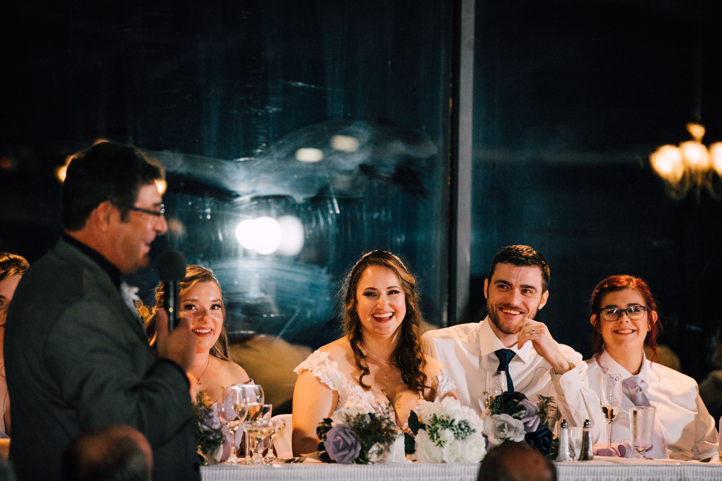  the bride and groom smile as a guest gives a toast during their wedding at endwell greens 