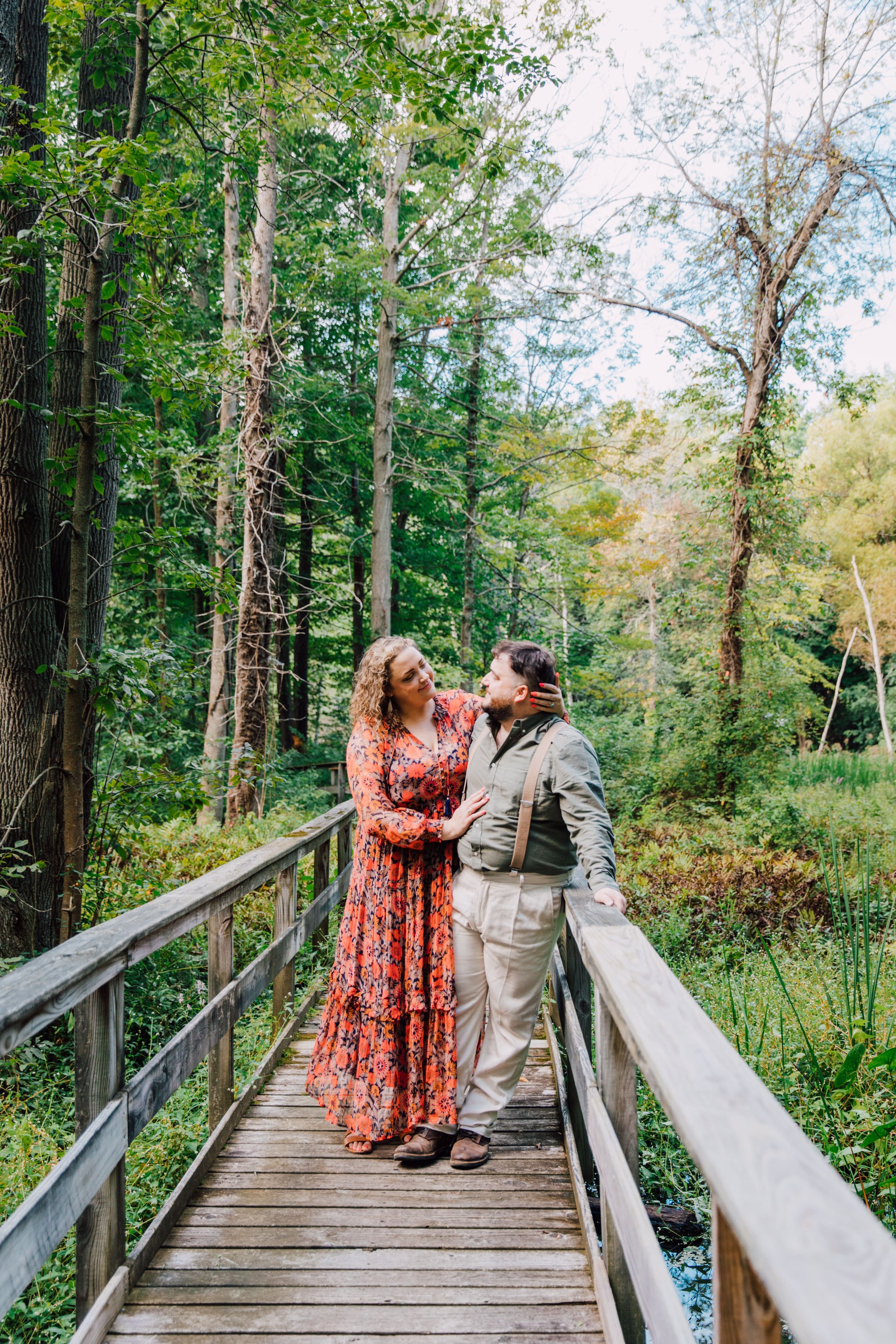  central new york photographer captures forest engagement photos for a couple as they stand on a wooden bridge over a forest area and smile at each other 