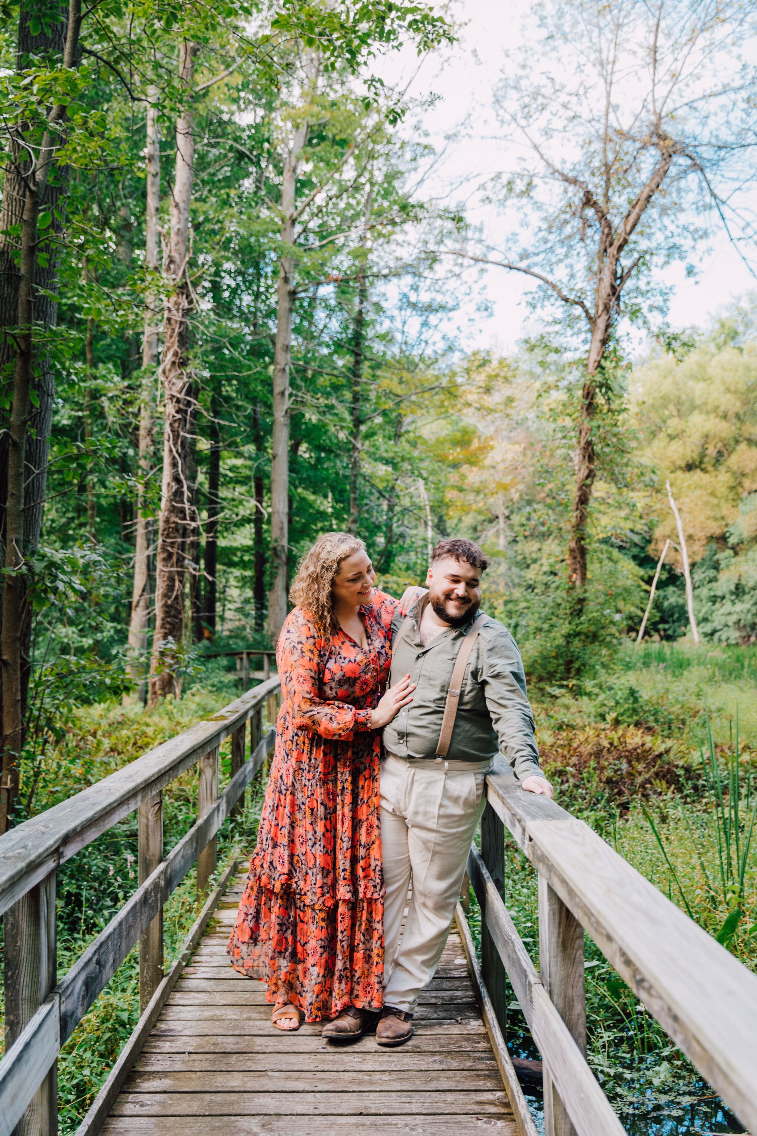  katie wraps her arms around her fiancee as they take engagement photos in the woods on a wooden bridge over a creek 
