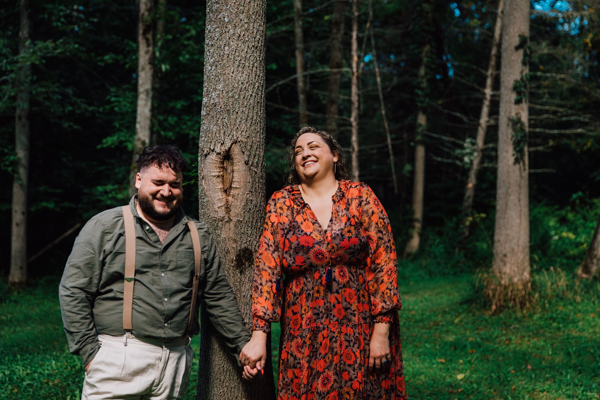  an engaged couple hold hands as they laugh and lean against a tree during their forest engagement photos 
