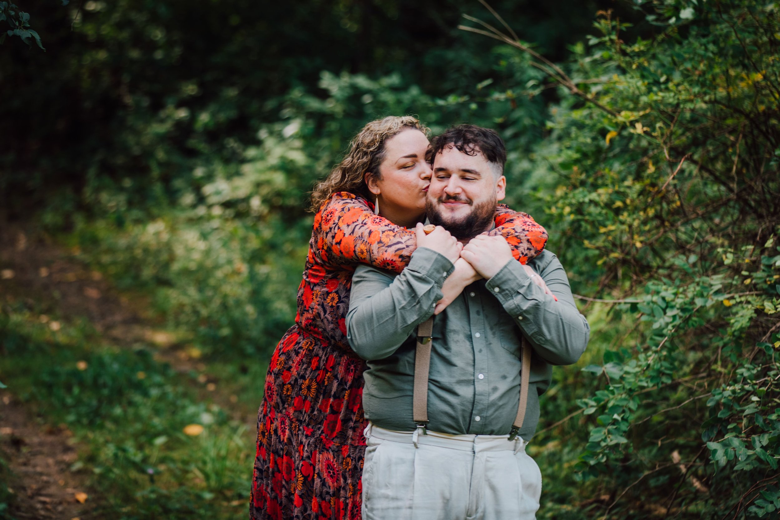  katie kisses her fiancee’s cheek as she stands behind him with her arms wrapped around his shoulders during their forest engagement photos 