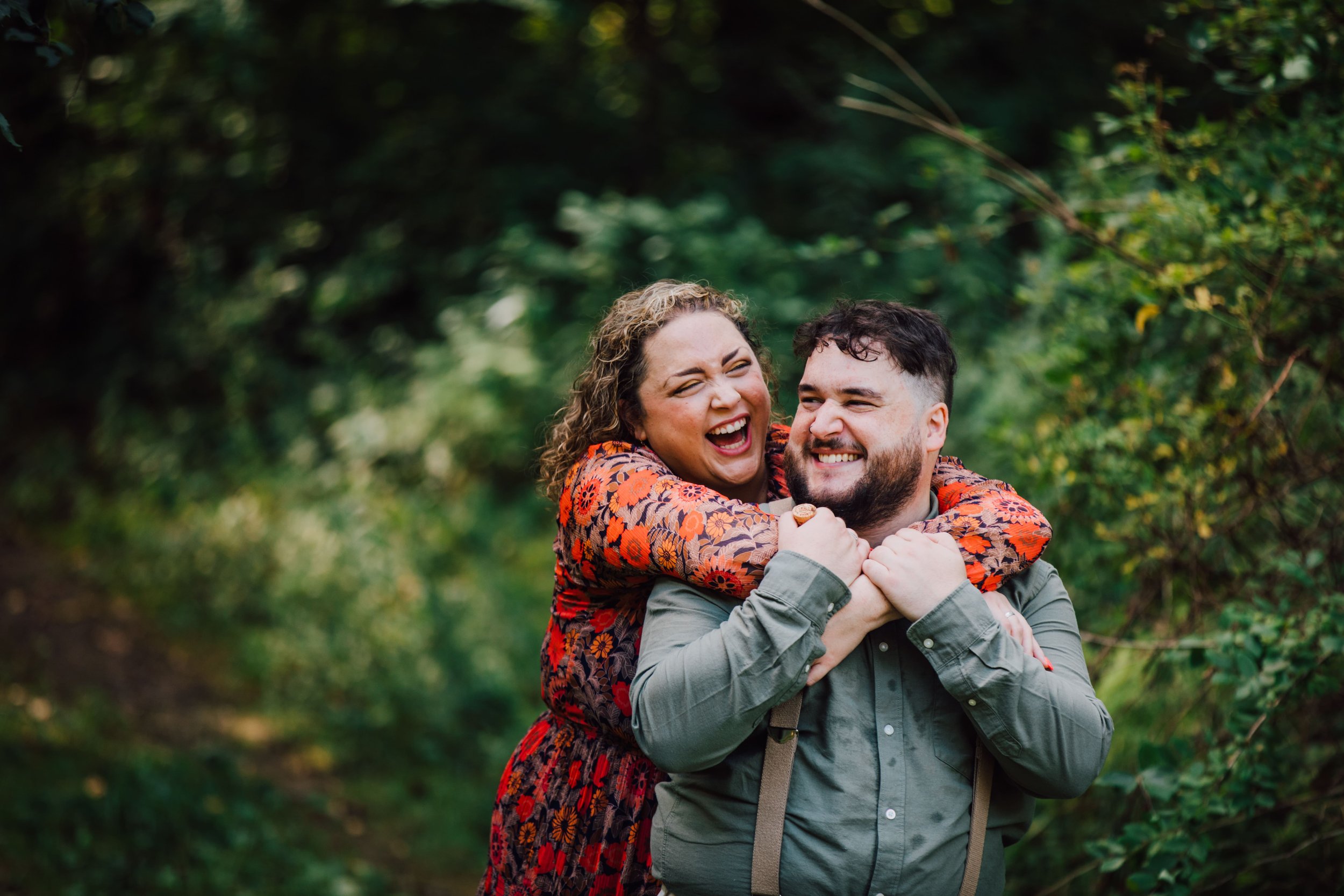  katie stands behind her fiancee with her arms wrapped around his shoulders as they laugh while taking engagement photos in the woods 