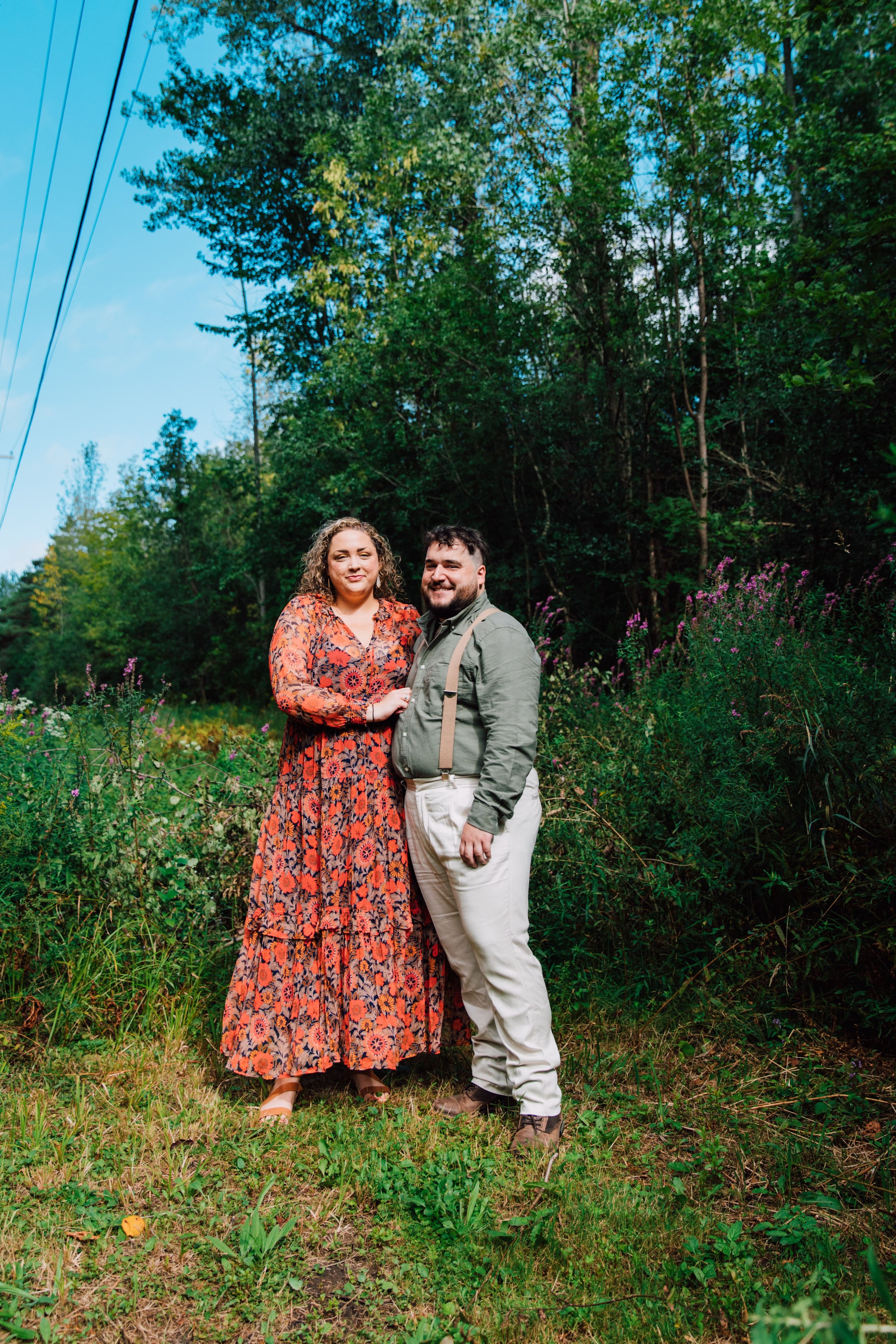  an engaged couple stand smiling in front of a field of wildflowers while taking engagement photos in the woods 