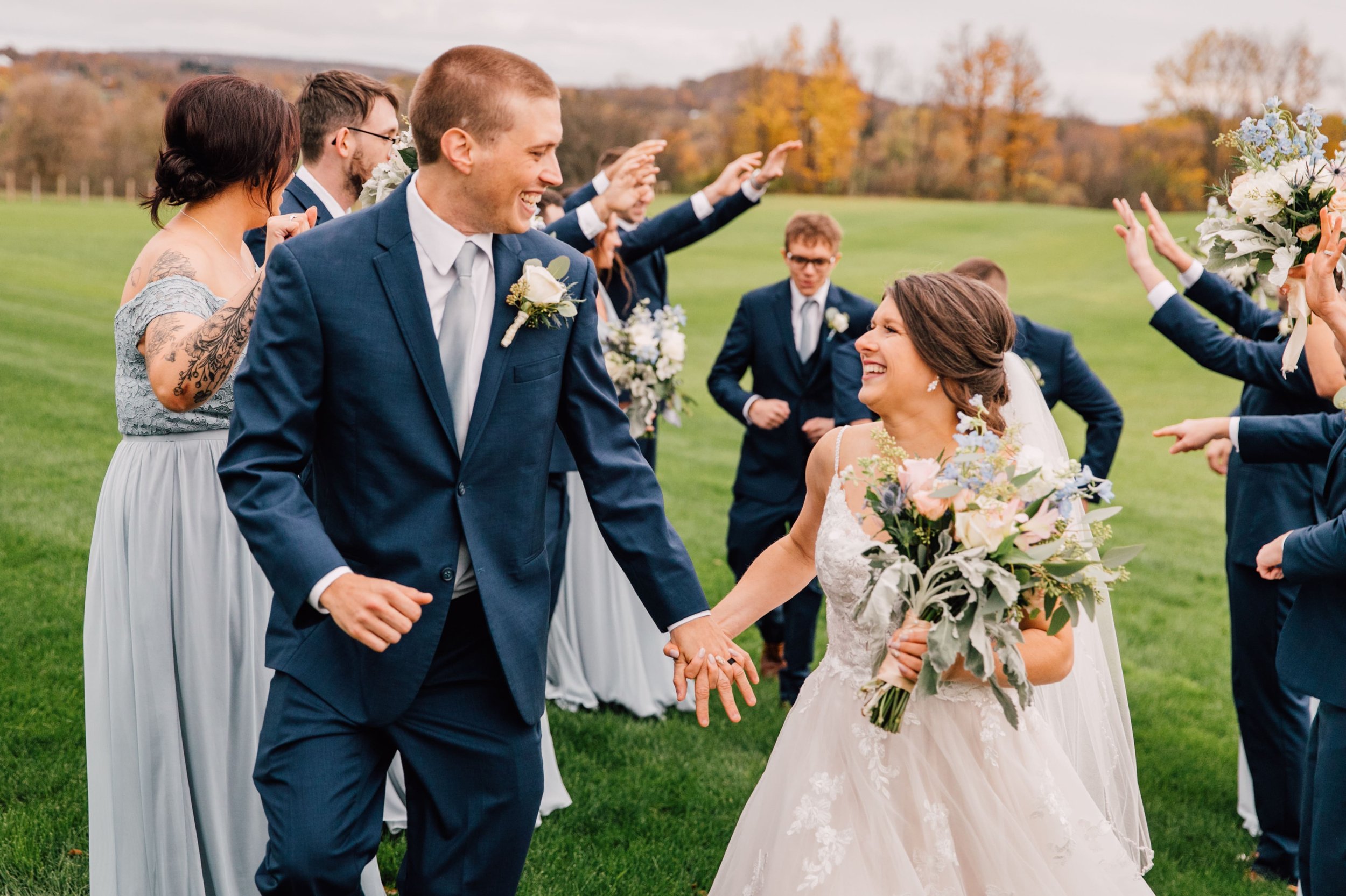  the bride and groom smile at each other while their bridal party creates an arch to run through at their outdoor fall wedding 