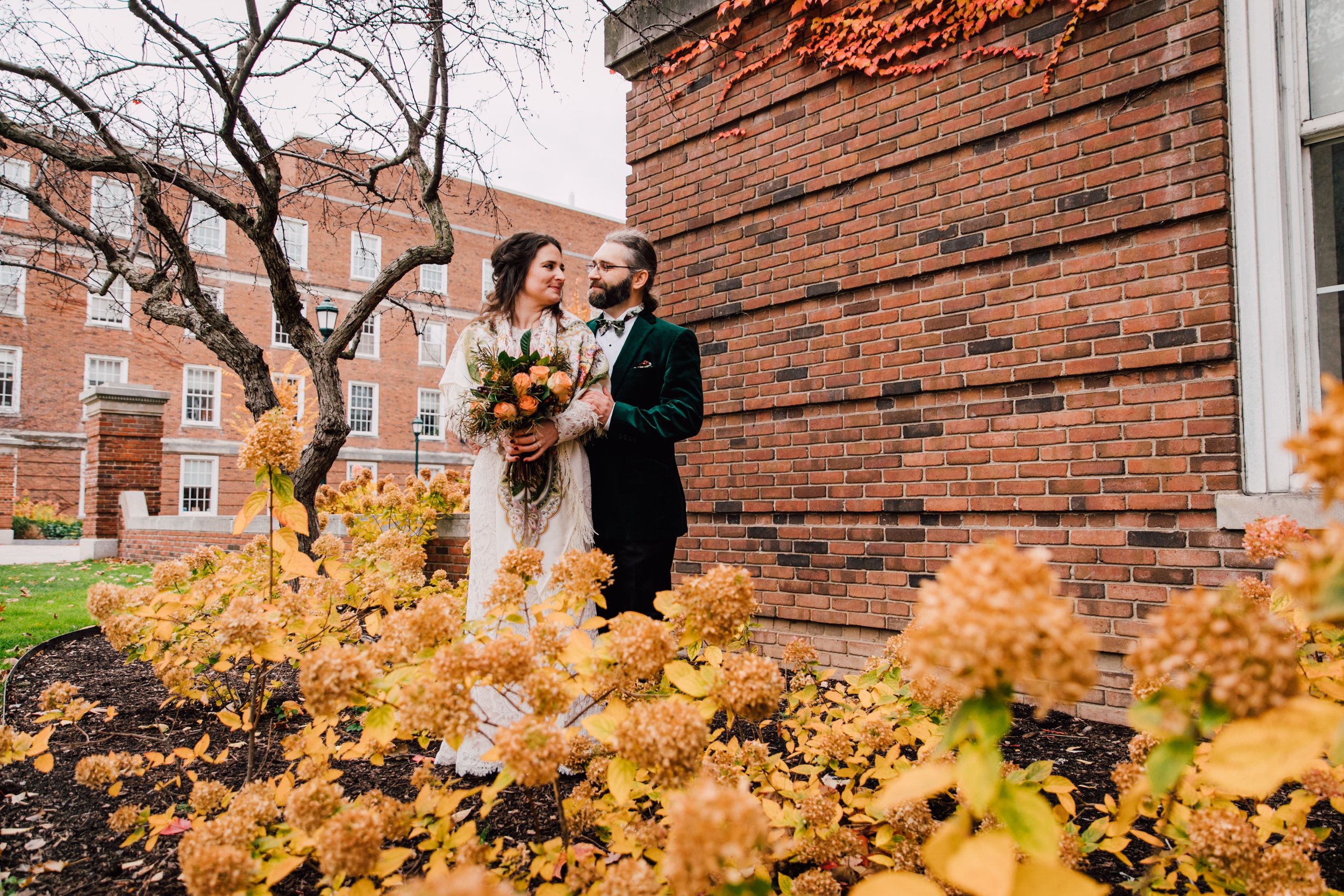 the bride and groom look at each other as the groom rests his hand on the brides arm and they stand in front of a brick building among fall foliage while syracuse ny wedding photographer captures their wedding portraits 