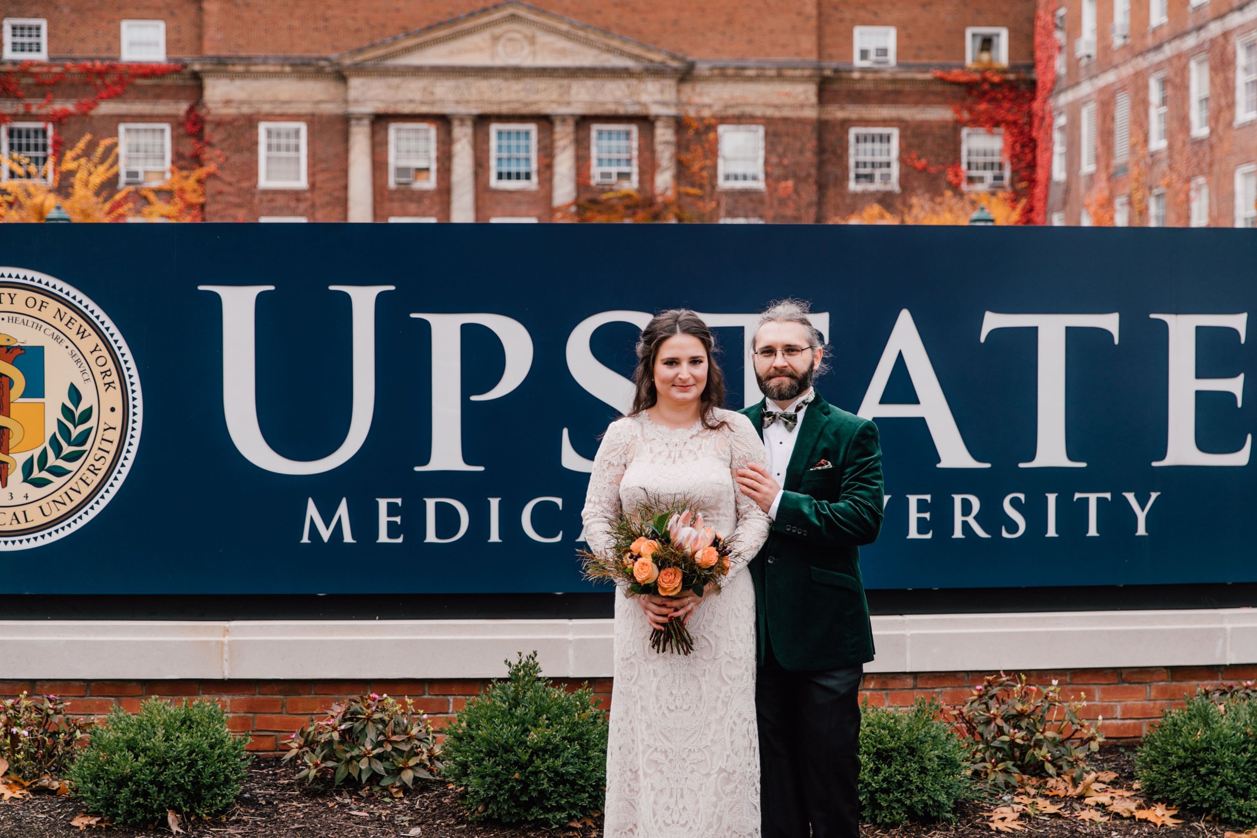  the bride and groom stand in front of the snyu upstate medical school sign, where they met. 