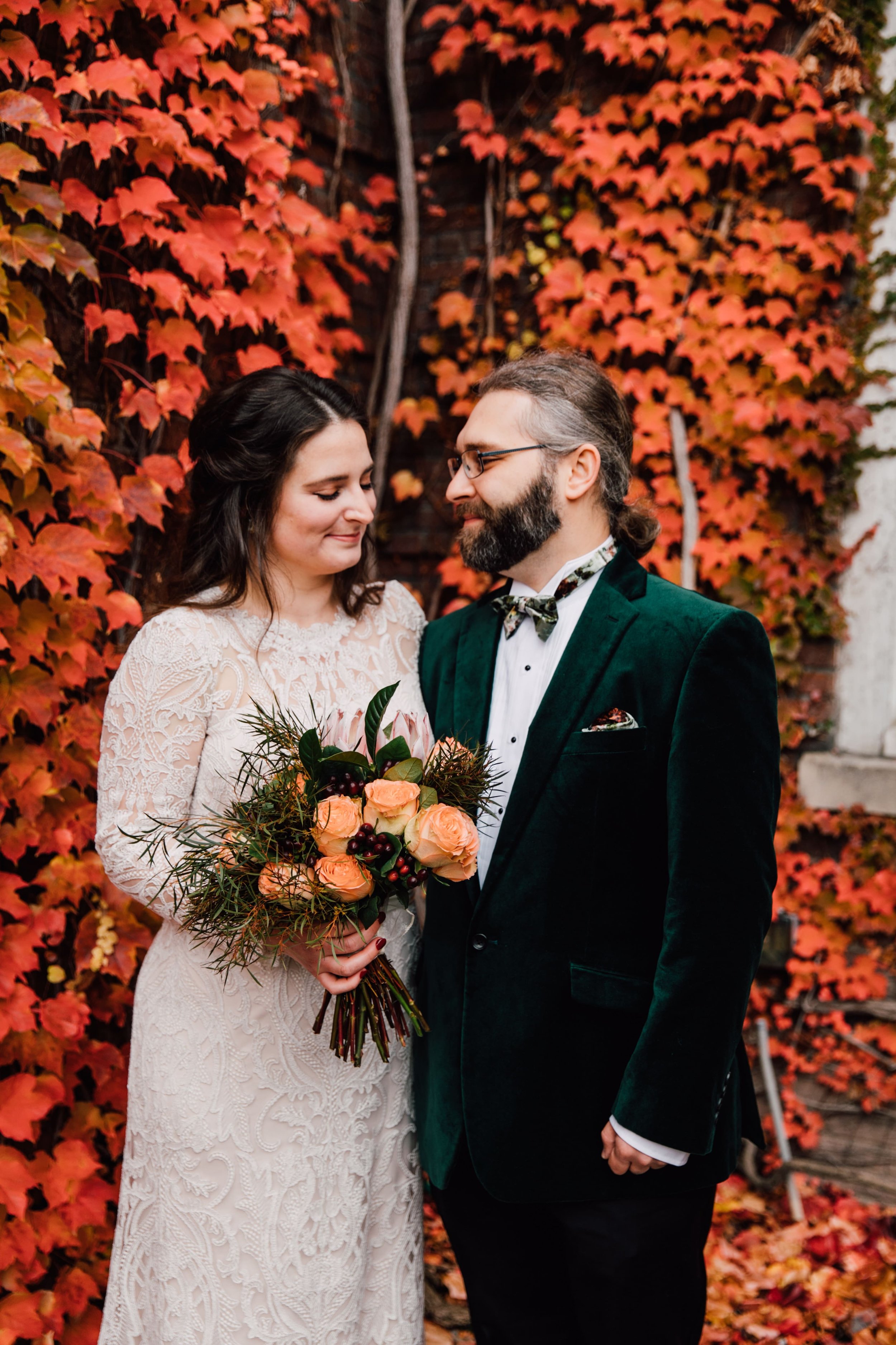  the bride and groom smile sweetly as they lean into each other and stand in front of fall orange ivy during wedding portrait session with syracuse ny wedding photographer 