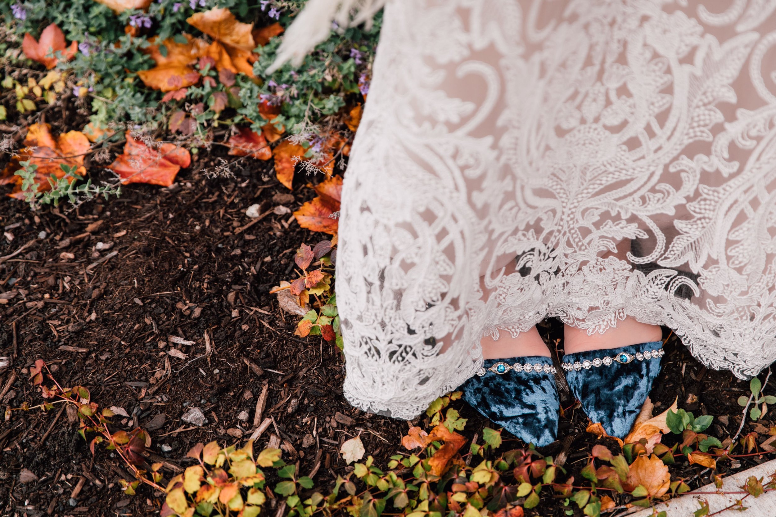  a detail shot captured by syracuse wedding photographer, the bride stands among fall leaves wearing blue velvet shoes with an eye detail. this closeup photo of her wedding shoes is special to them as they are scientists! 