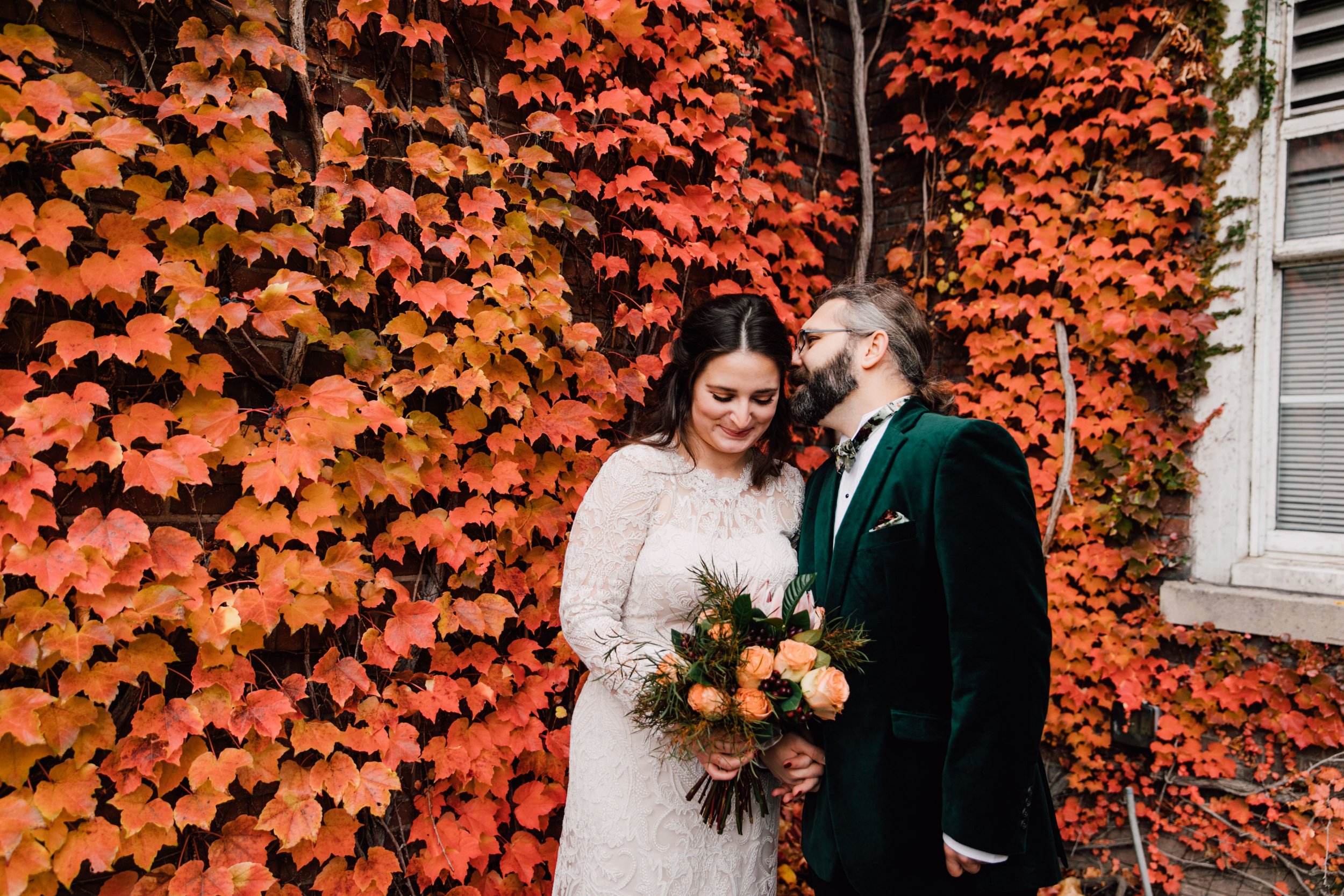  the groom whispers in the bride’s ear as she smiles and they stand in front of fall colored ivy during their syracuse ny wedding photography portrait session 