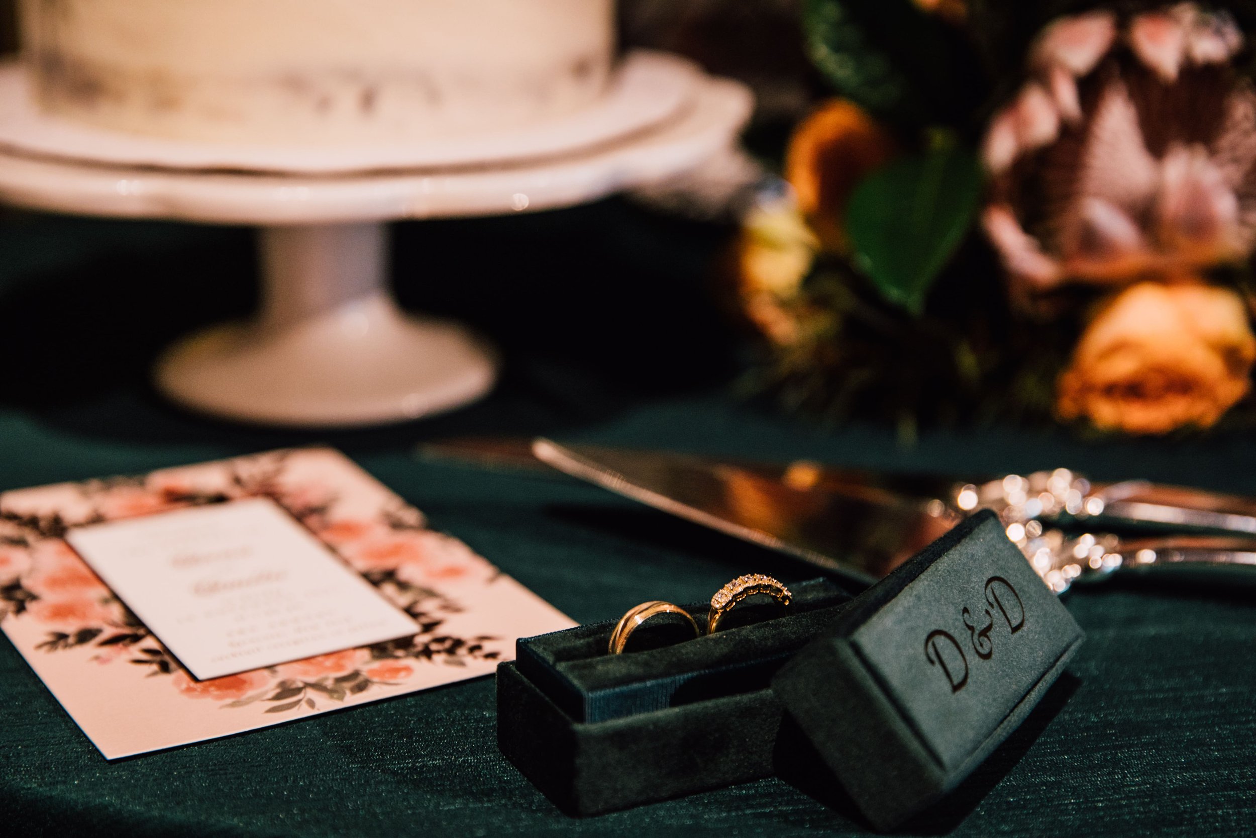  a syracuse wedding photographer snapped a beautiful photo of wedding day details, the couple’s rings sit in a green velvet jewelry box as their wedding cake, invitation, florals, and cake spatula rest out of focus in the background 
