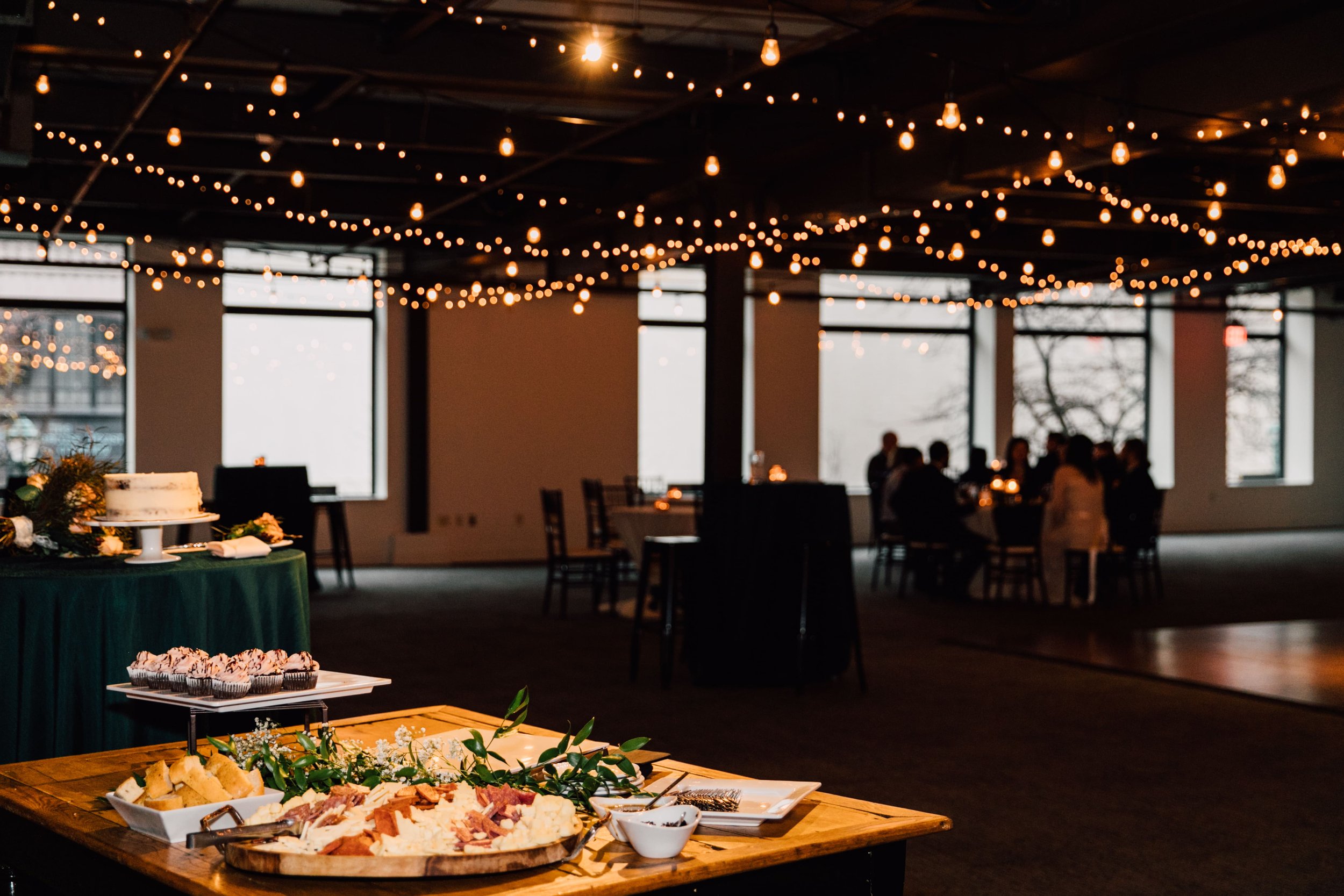  a photo from behind the food table at a sky armory wedding while guest silhouettes mingle in the background with string lights gleaming out of focus above them  