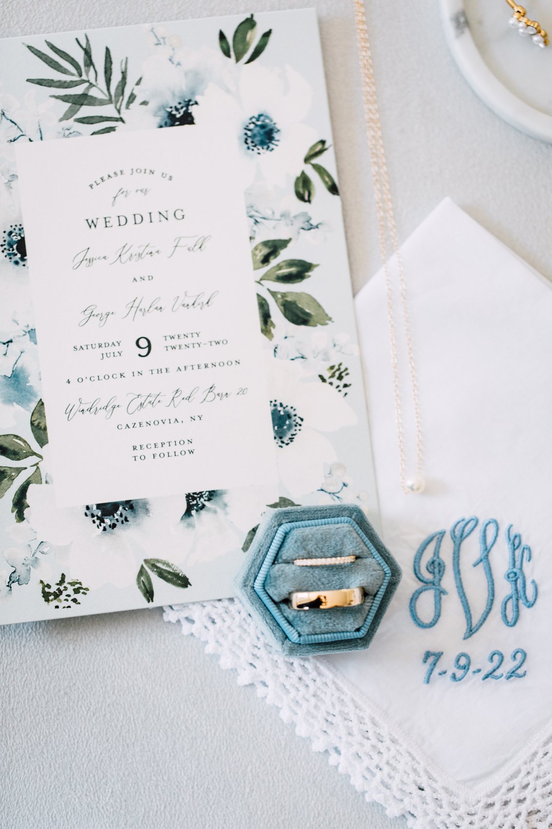  Layflat of a wedding invitation, embroidered handkerchief, necklace, and wedding rings with blue and white color scheme 