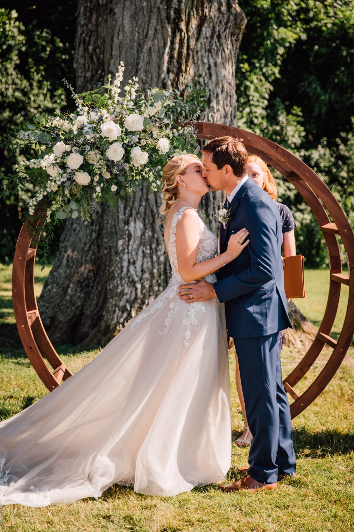  Bride and Groom share a first kiss at their outdoor wedding ceremony at Windridge Estate Red Barn, standing in front of a round arch with white roses and greenery 