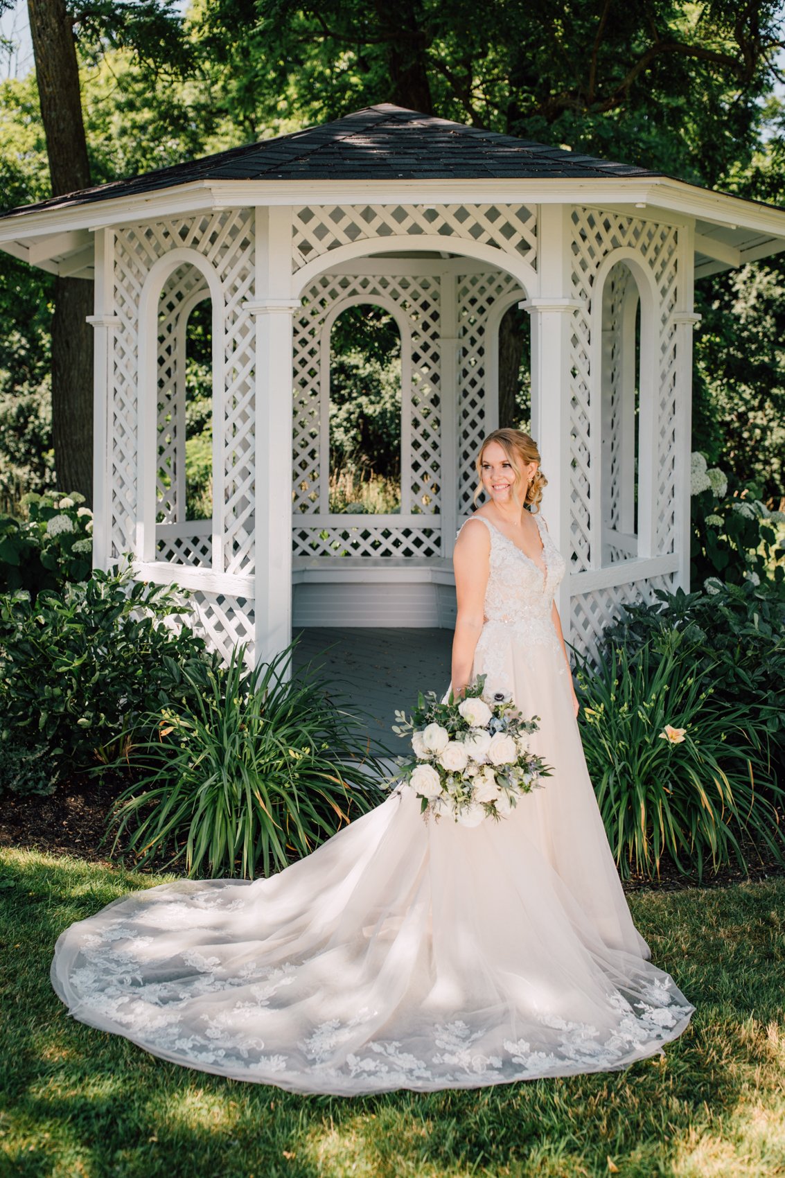  Bridal portrait in front of a white gazebo at Windridge Estate Red Barn in Central NY as the Bride wears a tulle dress with lace applique and holds a bouquet of white roses and greenery 