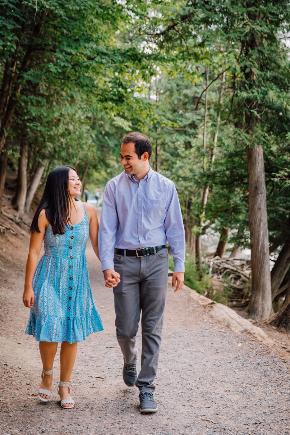  Engaged couple smiles and holds hands while walking along a path at Green Lakes State Park   
