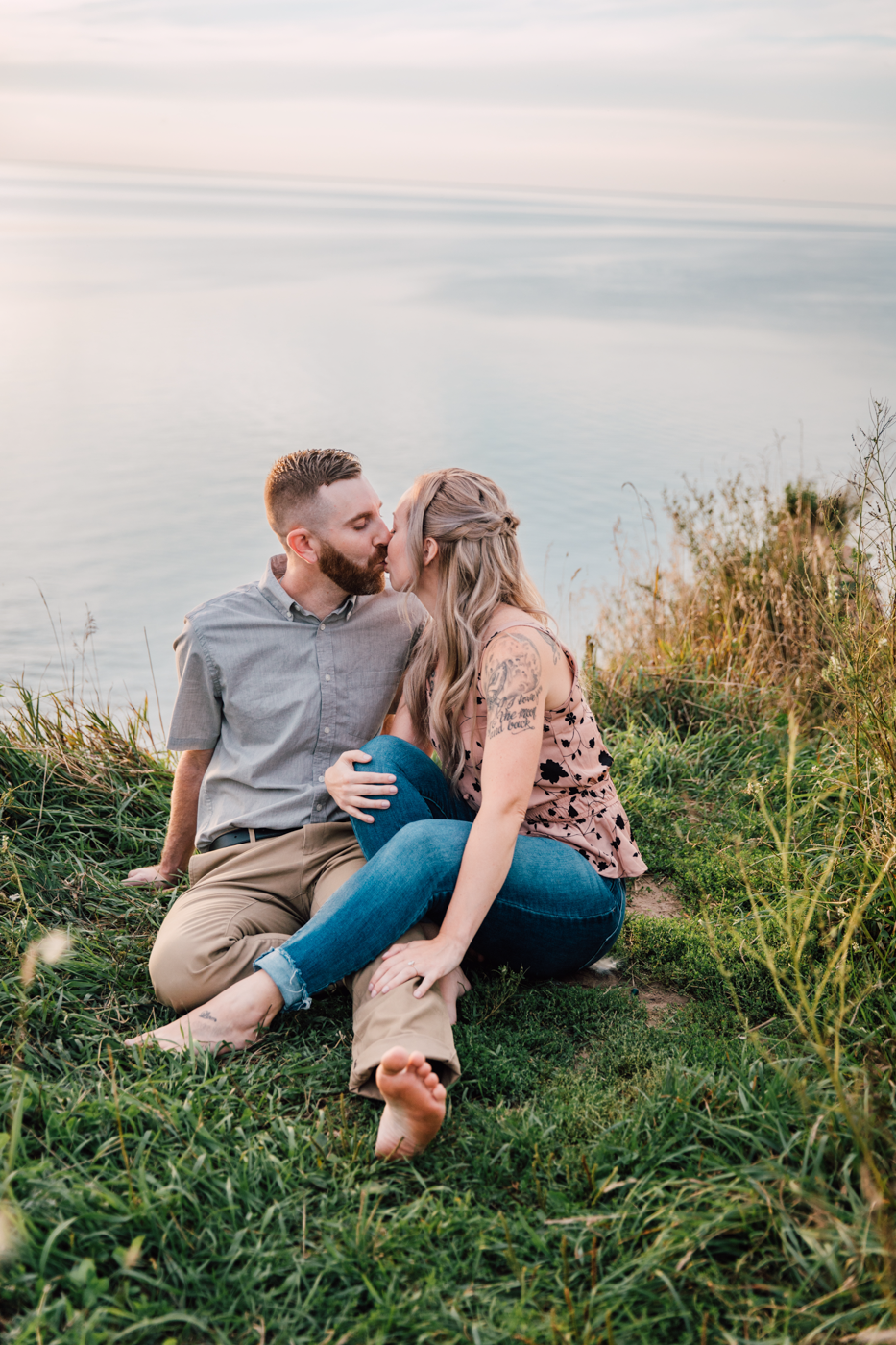  Engaged couple sits barefoot in the grass and kisses, along the Oswego bluffs in central NY 