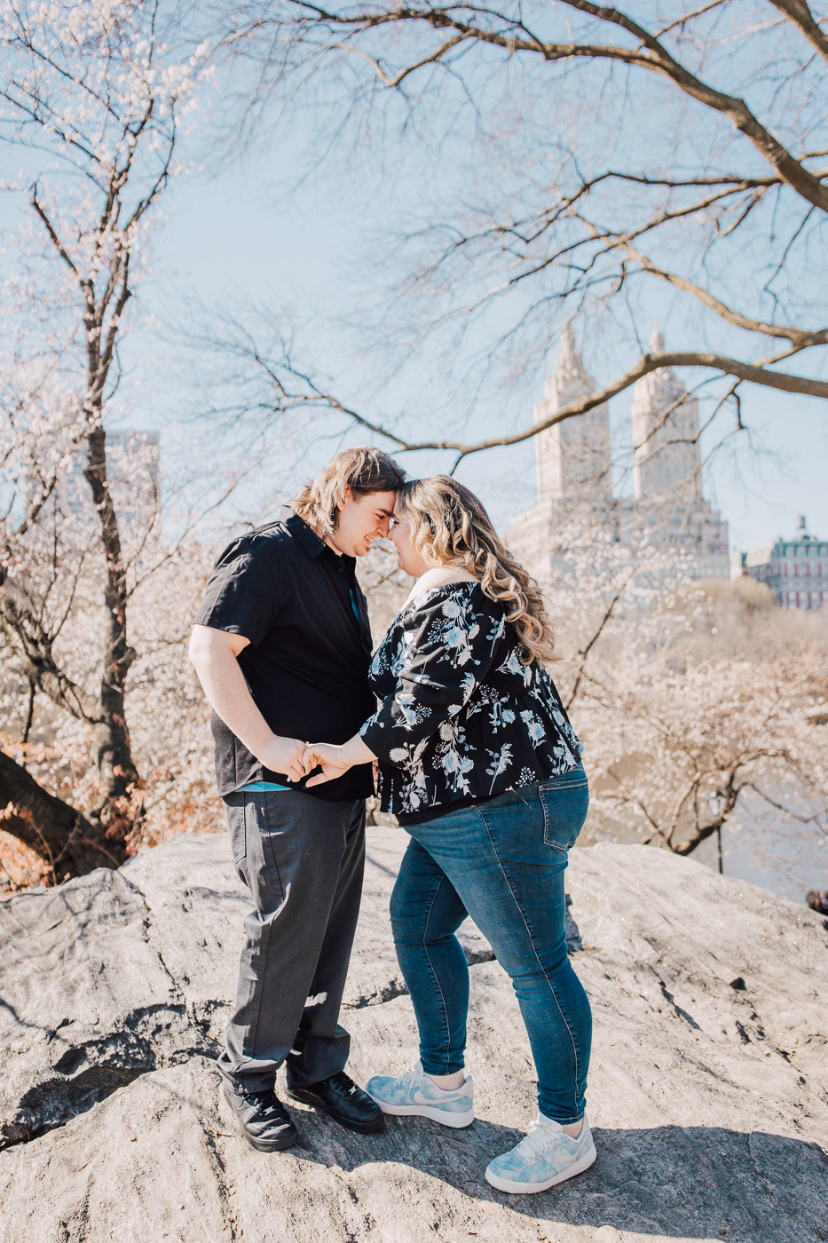  Engaged couple holds hands with the New York City skyline behind them while taking Central Park engagement photos 