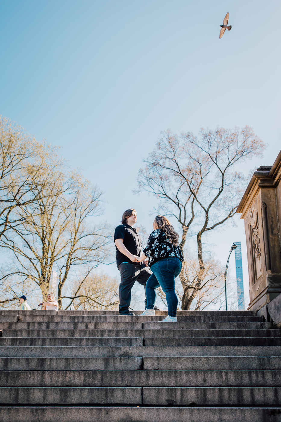  engaged couple stands atop the Bethesda Terrace steps in Central Park in spring while taking New York City engagement photos   