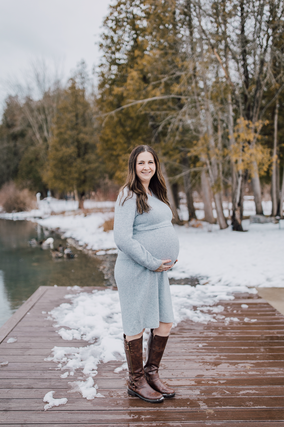  Pregnant woman holds her belly while standing on a dock at Green Lakes State Park during her winter photo shoot 