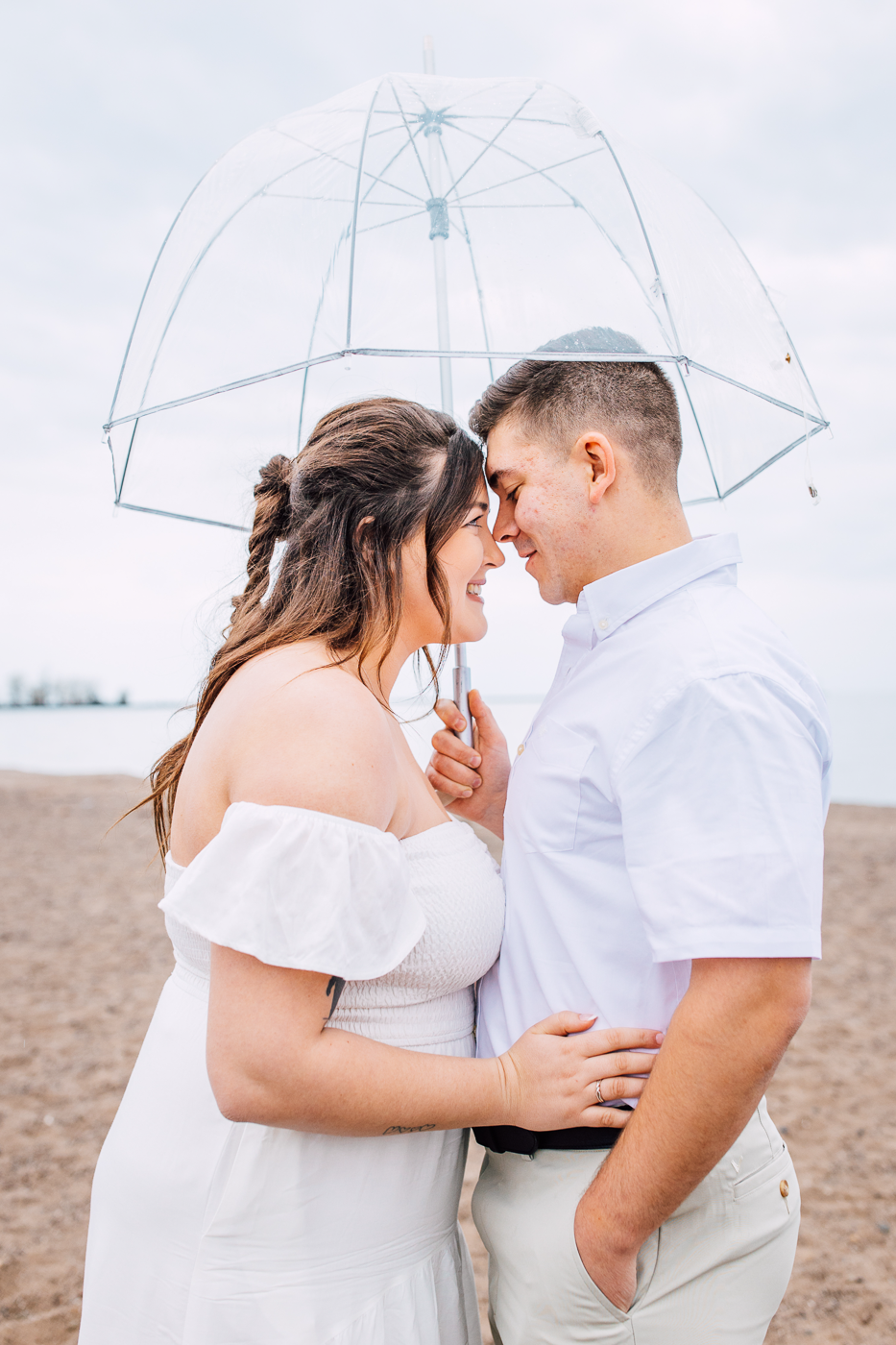  rainy photoshoot on a lake ontario beach in central NY. Engaged couple cuddles up under an umbrella on a cloudy day on a the beach. 