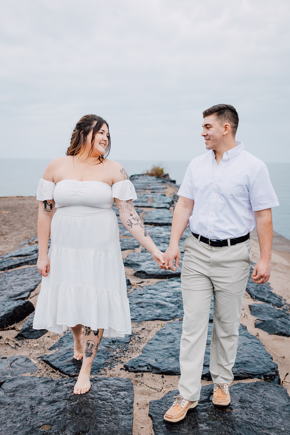  Engaged couple smiles at each other while holding hands on the Lake Ontario beach while Brittany Juravich takes some lakefront photography 