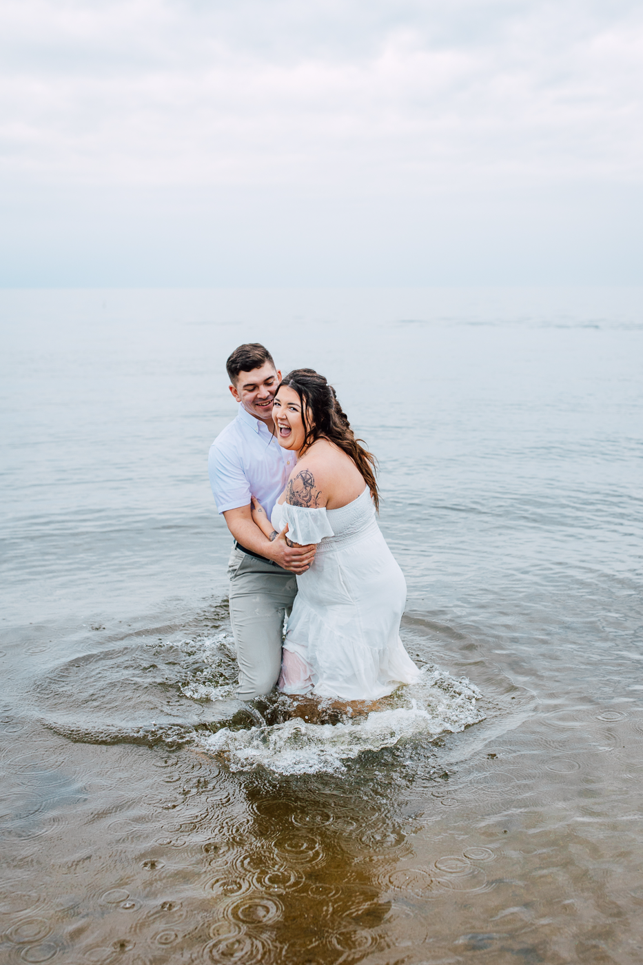  Woman laughs with her fiance as they stand in the water of Lake Ontario during their beach engagement photos at Fair Haven State Park 