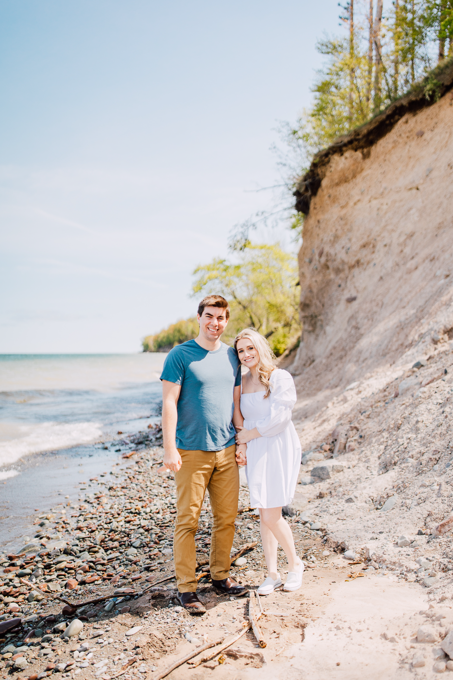  Engaged couple stands below Sterling Bluffs along Lake Ontario while taking spring engagement photos with Brittany Juravich 