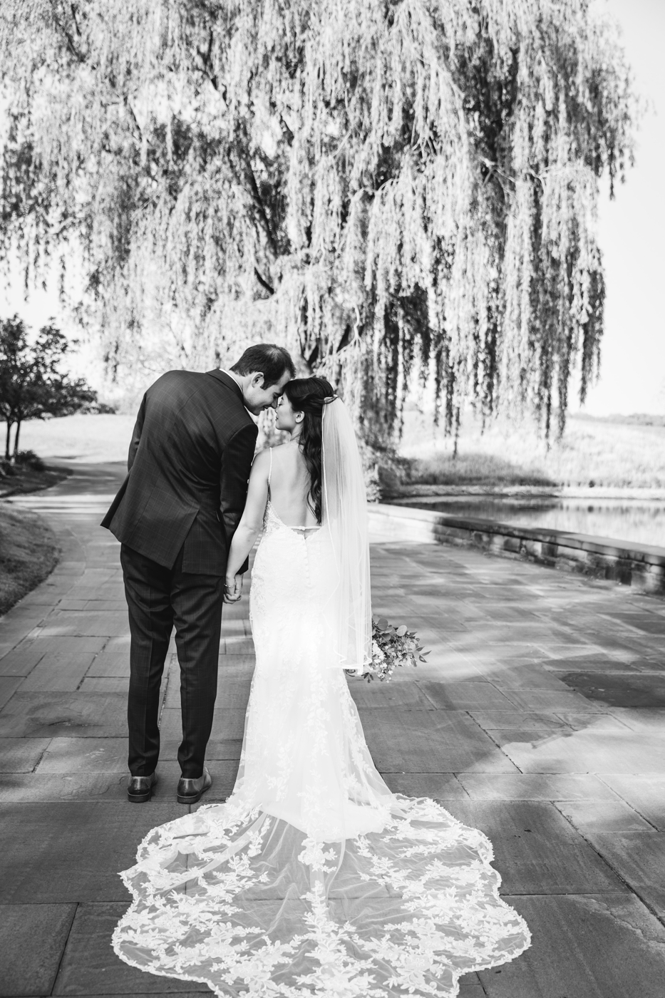  Black and white photo of a Bride and Groom with their backs to the camera and the Bride’s beautiful lace train on display during their elegant wedding at Lodge at Turning Stone 