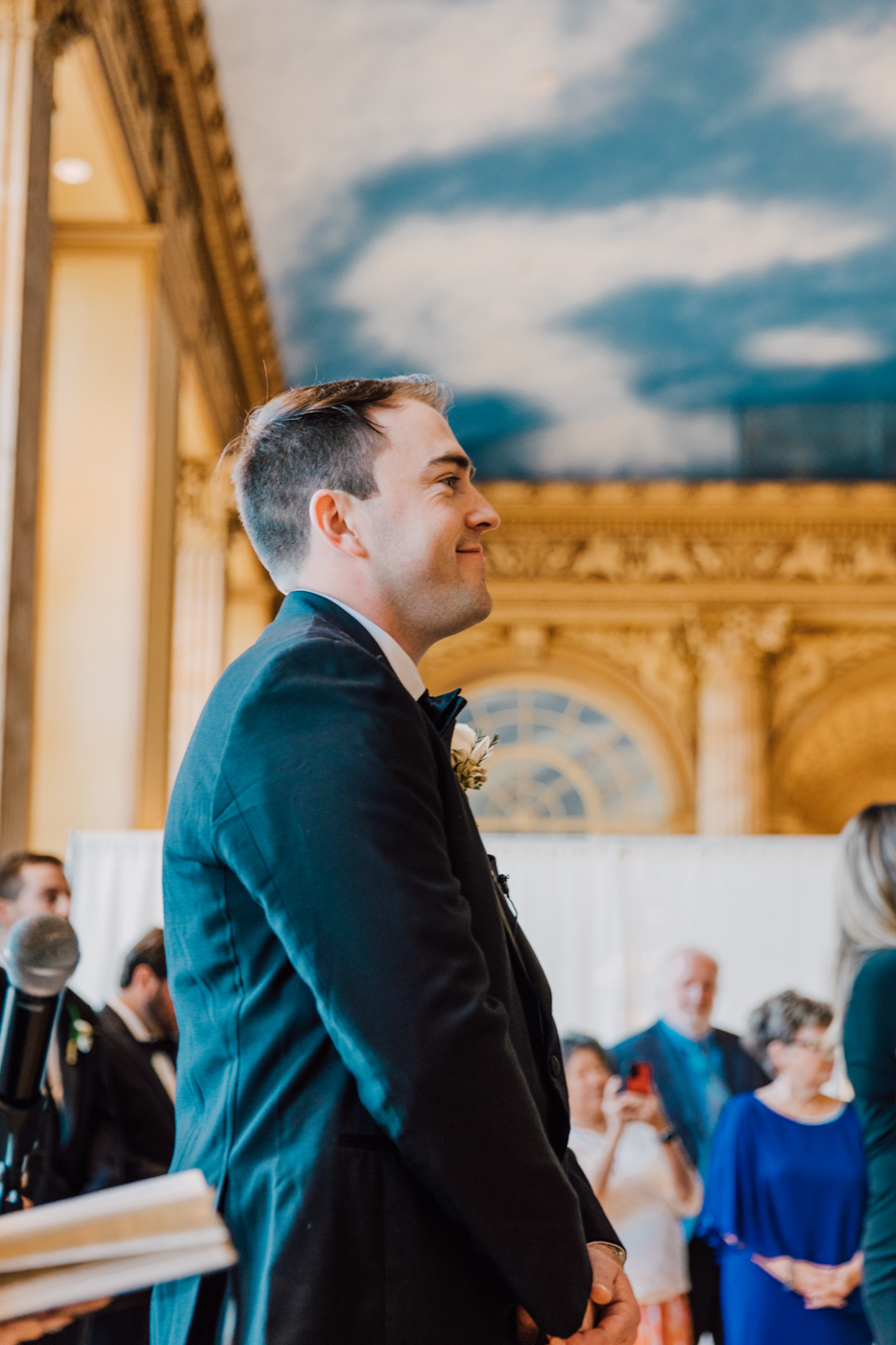  Groom smiles as he watches his Bride walk down the aisle toward him during their traditional wedding at the Marriott Syracuse Downtown 