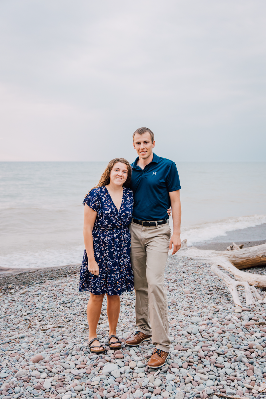  Engaged couple stand together on the rocky Lake Ontario beach during their summer engagement photos in Central NY 