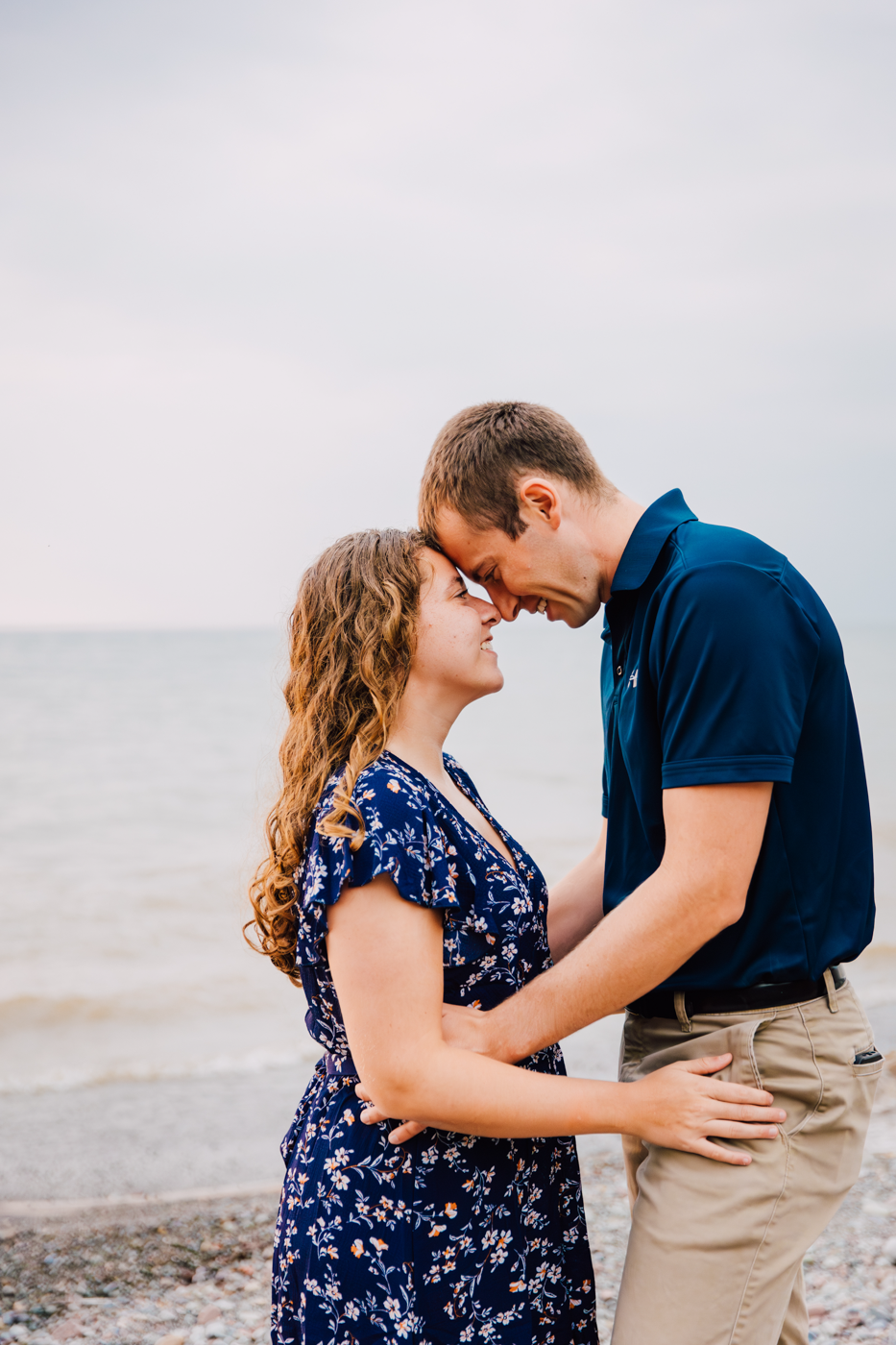  Engaged couple stands face-to-face during their engagement session with Brittany Juravich on the shores of Lake Ontario 