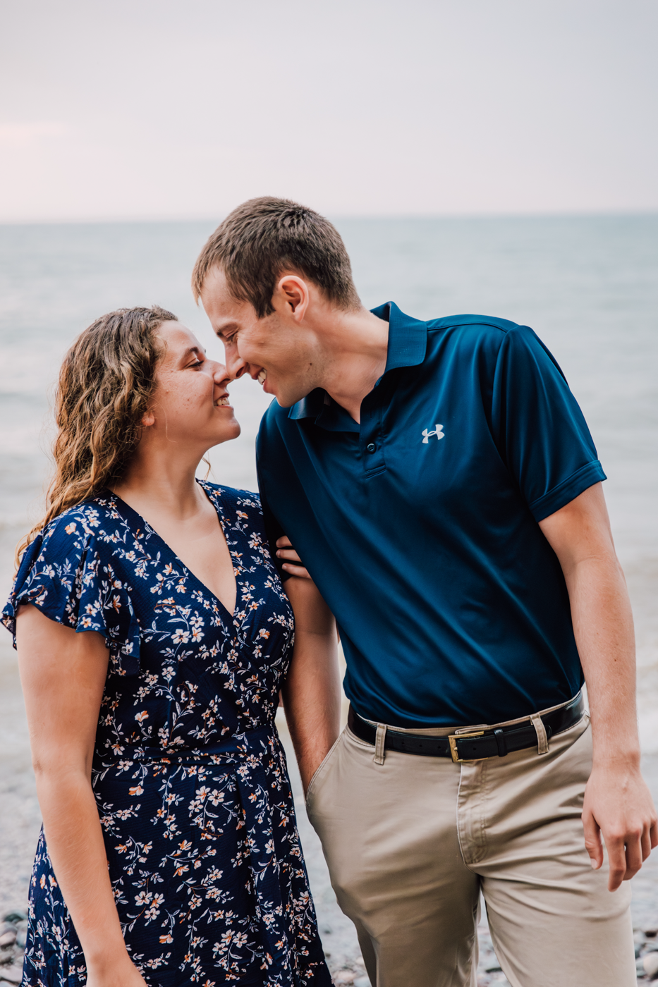  Engaged couple stand nose-to-nose in front of Lake Ontario during their summer engagement photos at the bluffs in Central NY 