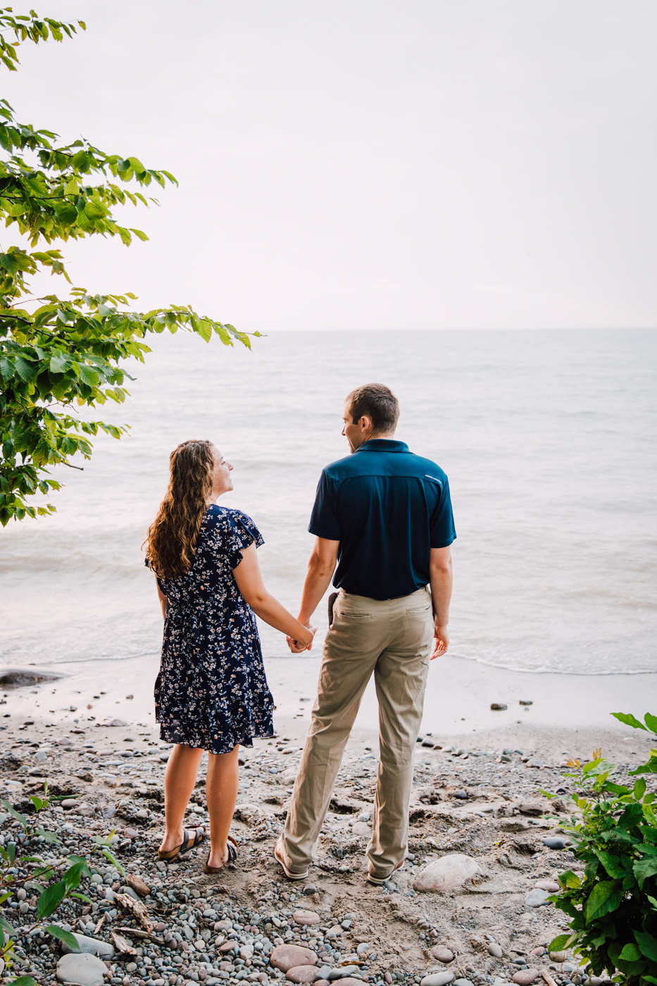  Engaged couple hold hands on the Lake Ontario beach while looking out at the water during their summer engagement photos 