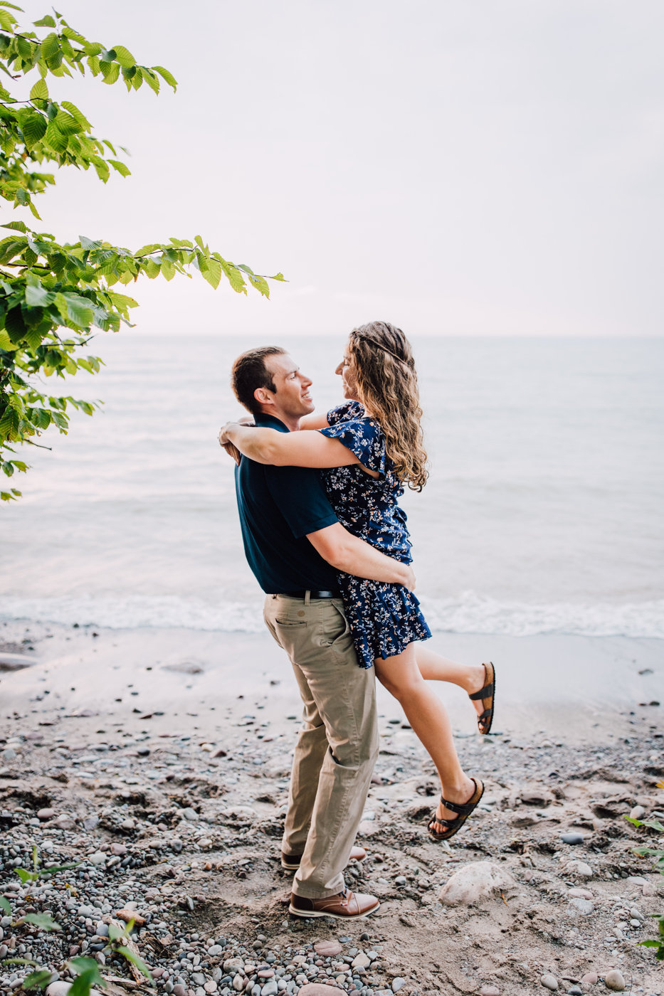  Man lifts his fiance up while standing on the Lake Ontario beach for their summer engagement photos with Brittany Juravich 