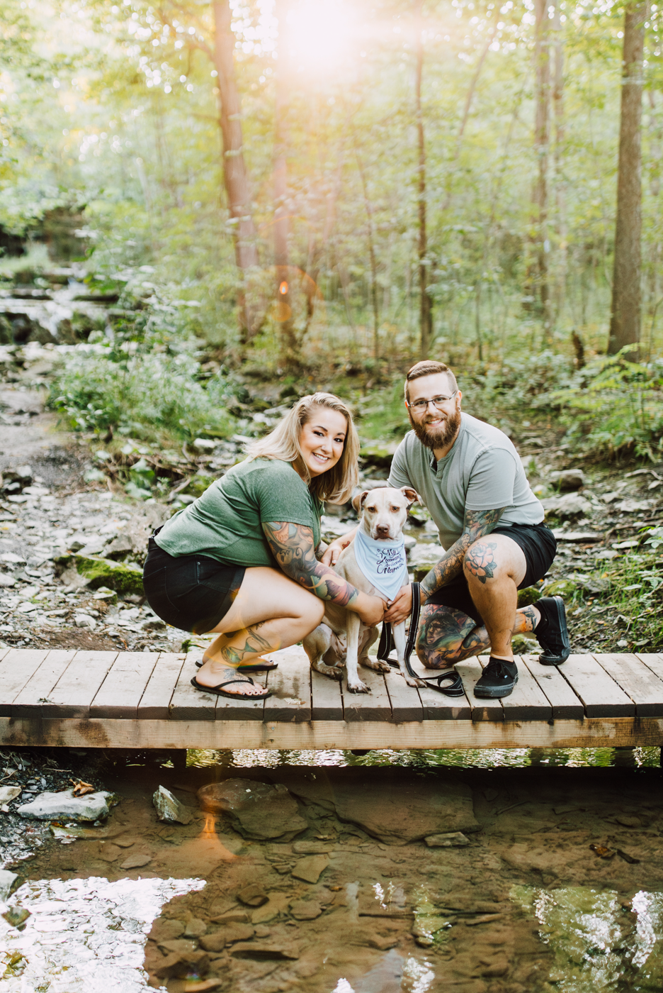  Engaged couple crouches with their dog on a small wooden bridge over a creek at Three Falls Woods during their forest photoshoot 