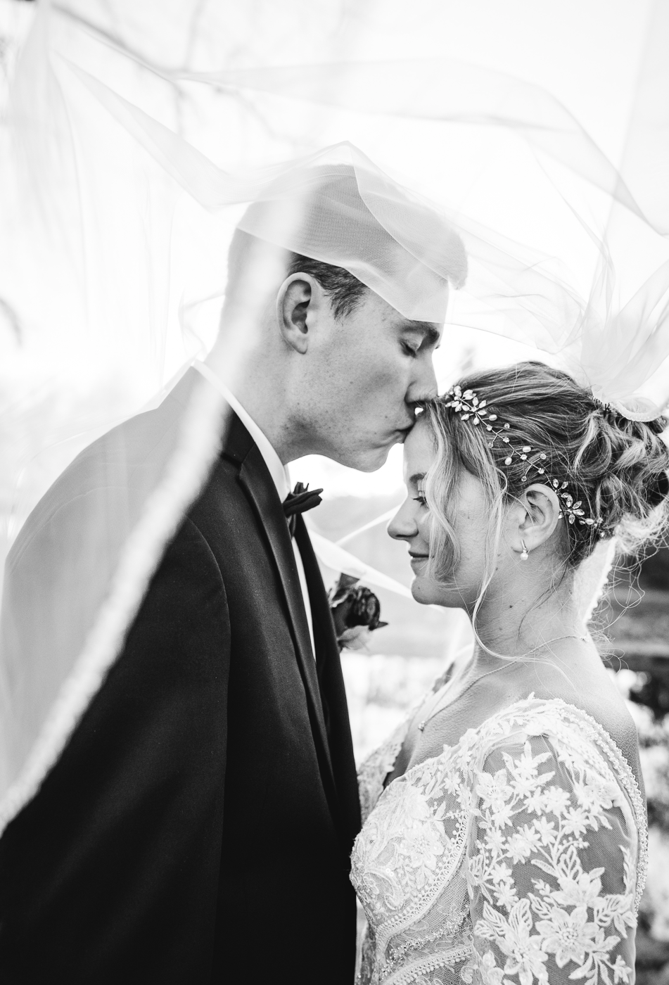  Black and white photo of Groom kissing his Bride on the forehead underneath her veil 