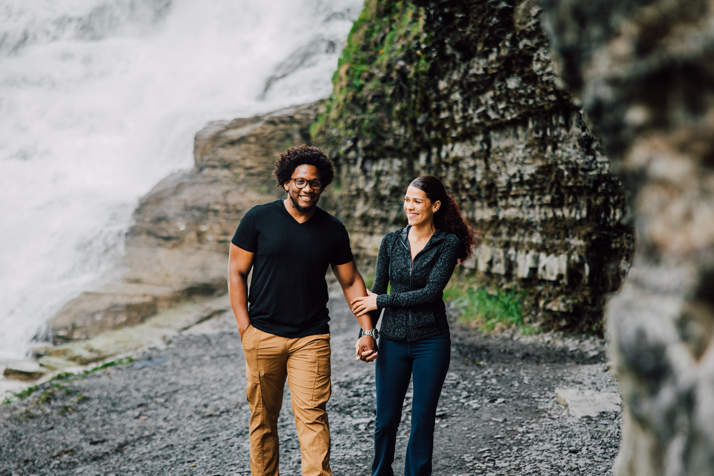Engaged couple hold hands as they walk near a waterfall during their proposal pictures at Ithaca Falls