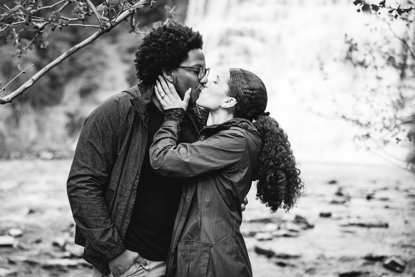 Black and white image of a newly engaged couple kissing in front of a waterfall