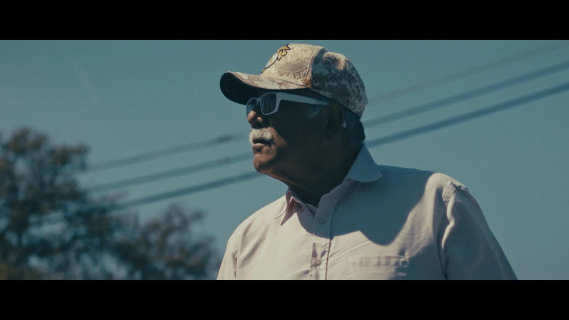 An older man with gray hair and mustache wearing a light-colored button-up shirt, dark sunglasses, and a camouflage baseball cap, standing outdoors against a clear blue sky with power lines in the background.