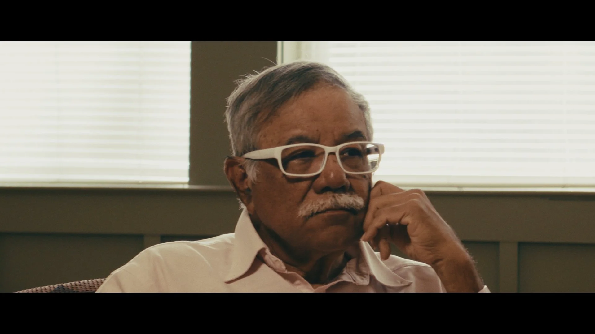 An elderly man with gray hair and a mustache, wearing white-rimmed glasses, is sitting indoors near windows with blinds, resting his chin on his hand and looking contemplative.