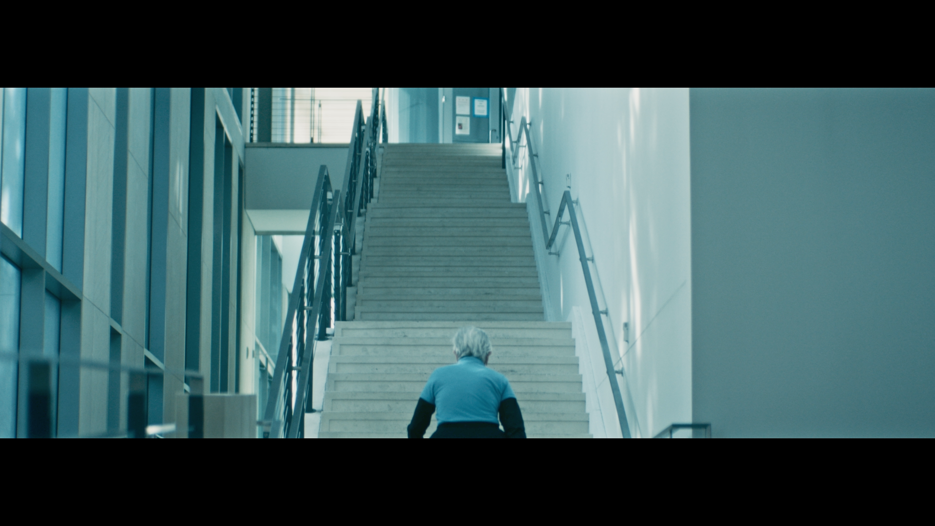 A person with gray hair wearing a light blue shirt walking up a staircase inside a modern building with glass walls and metal railings.
