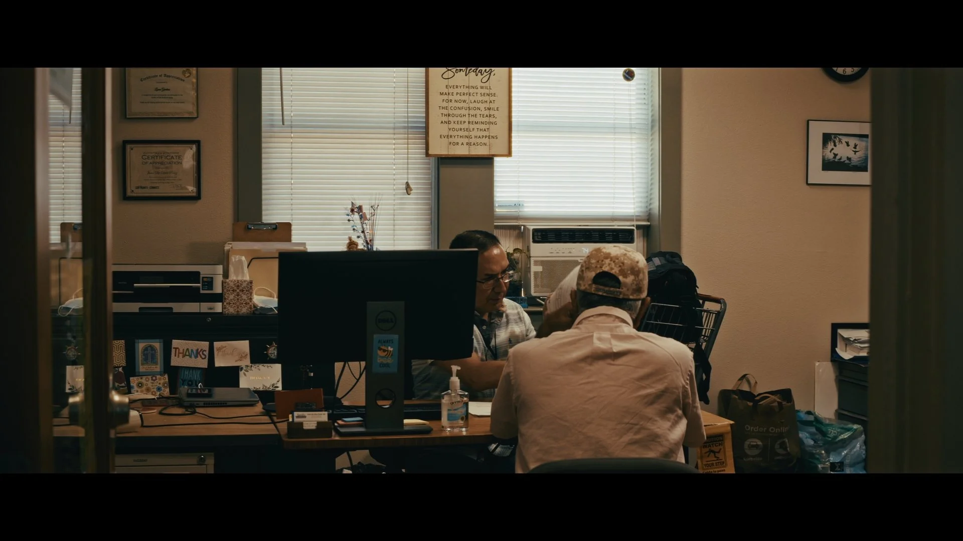 Two men are seated in an office, engaged in conversation. The room has a desk with a computer, decorations, and posters on the wall. Behind them, there are windows with blinds, an air conditioning unit, and various office supplies and bags.