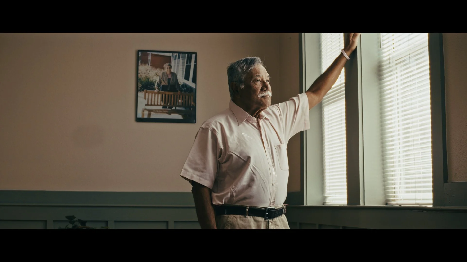 An elderly man stands by a window with blinds, looking outside with a contemplative expression, wearing a light pink short-sleeved shirt and khaki pants.
