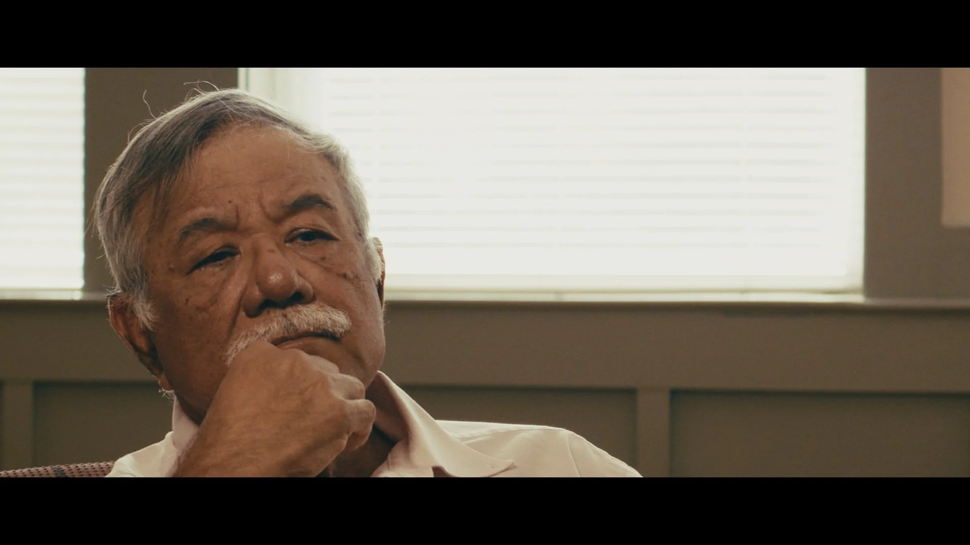 An elderly man with gray hair, a mustache, and brown skin is sitting indoors near a window with blinds, resting his chin on his hand and looking thoughtful.