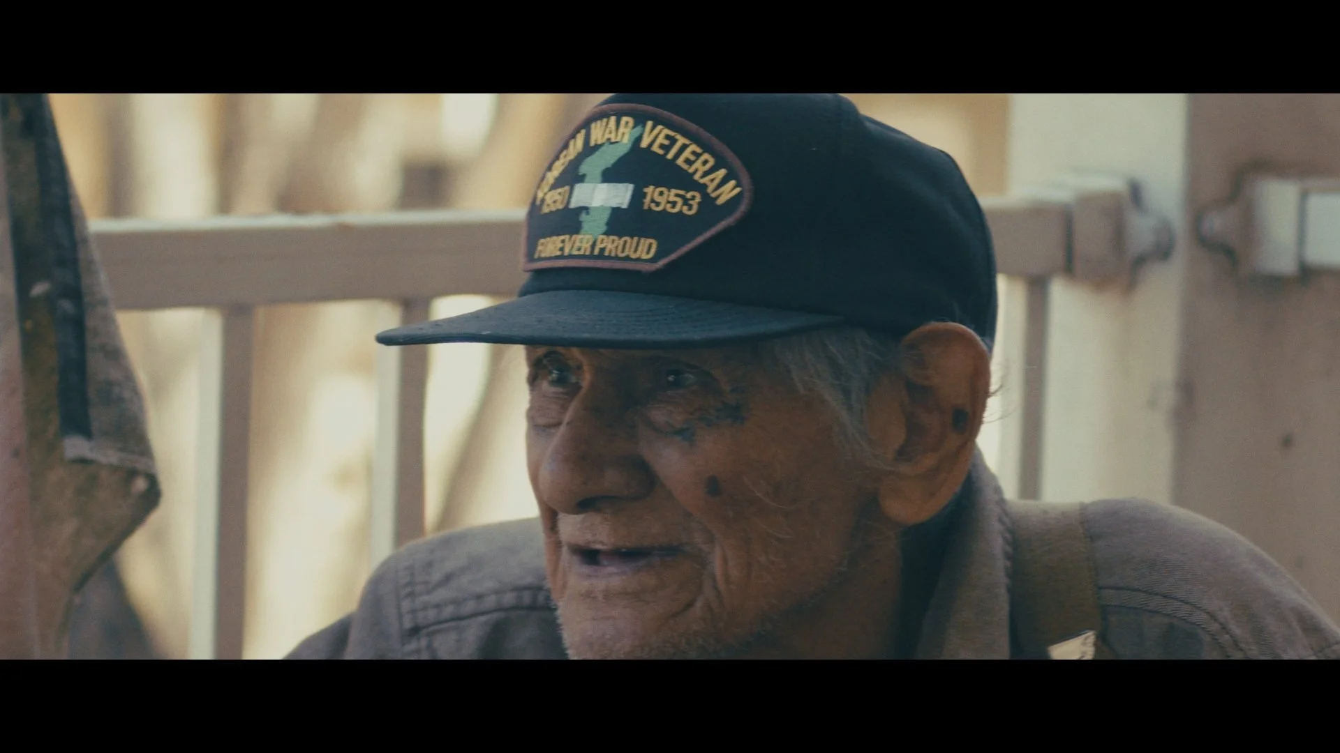 An elderly man with a weathered face and wrinkles, wearing a black Veteran cap with an American War Veteran patch, is sitting outdoors near a fence.