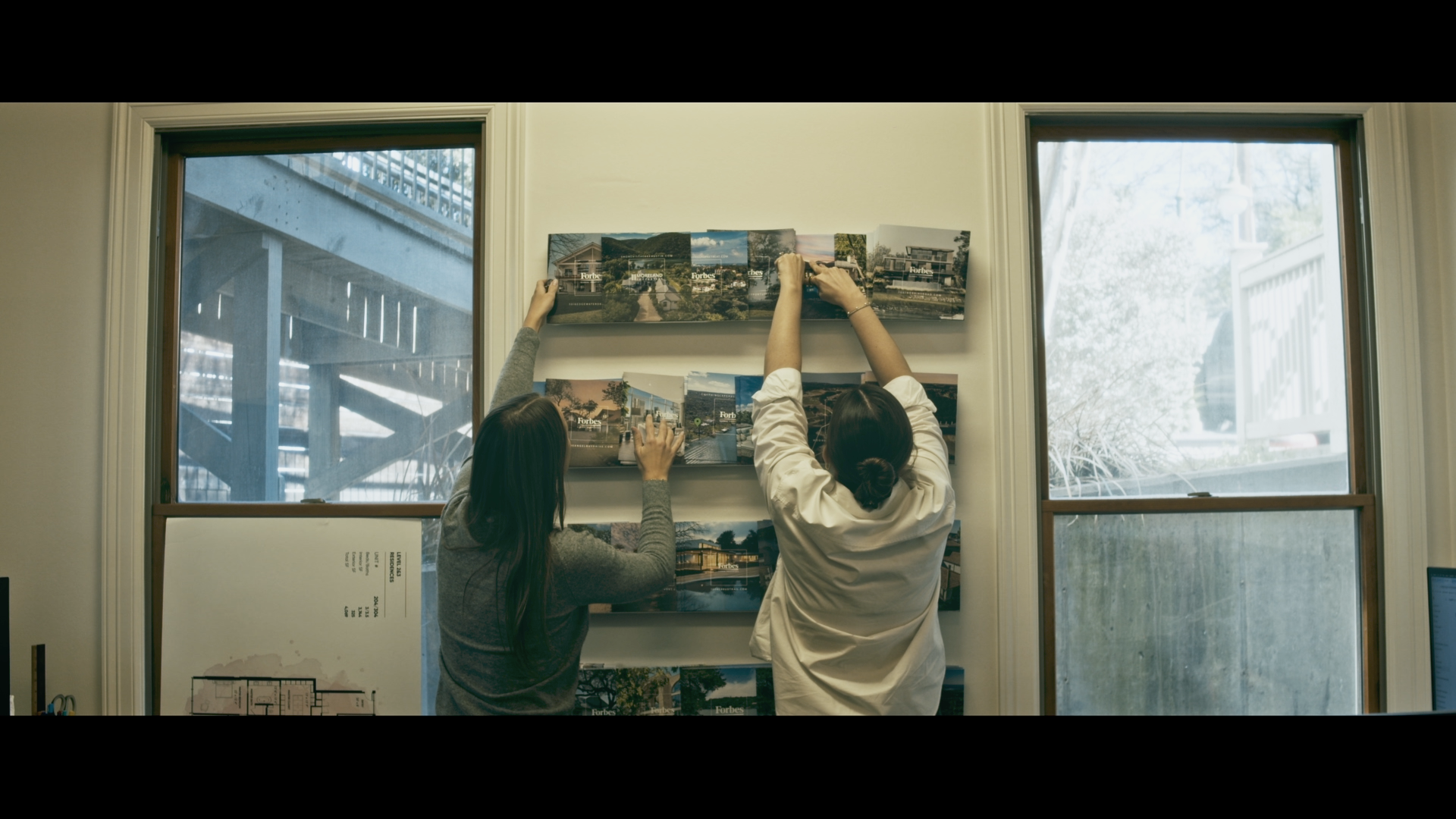 Two women hang photographs on a white wall inside a room with large windows. Outside the windows, building structures and trees are visible.