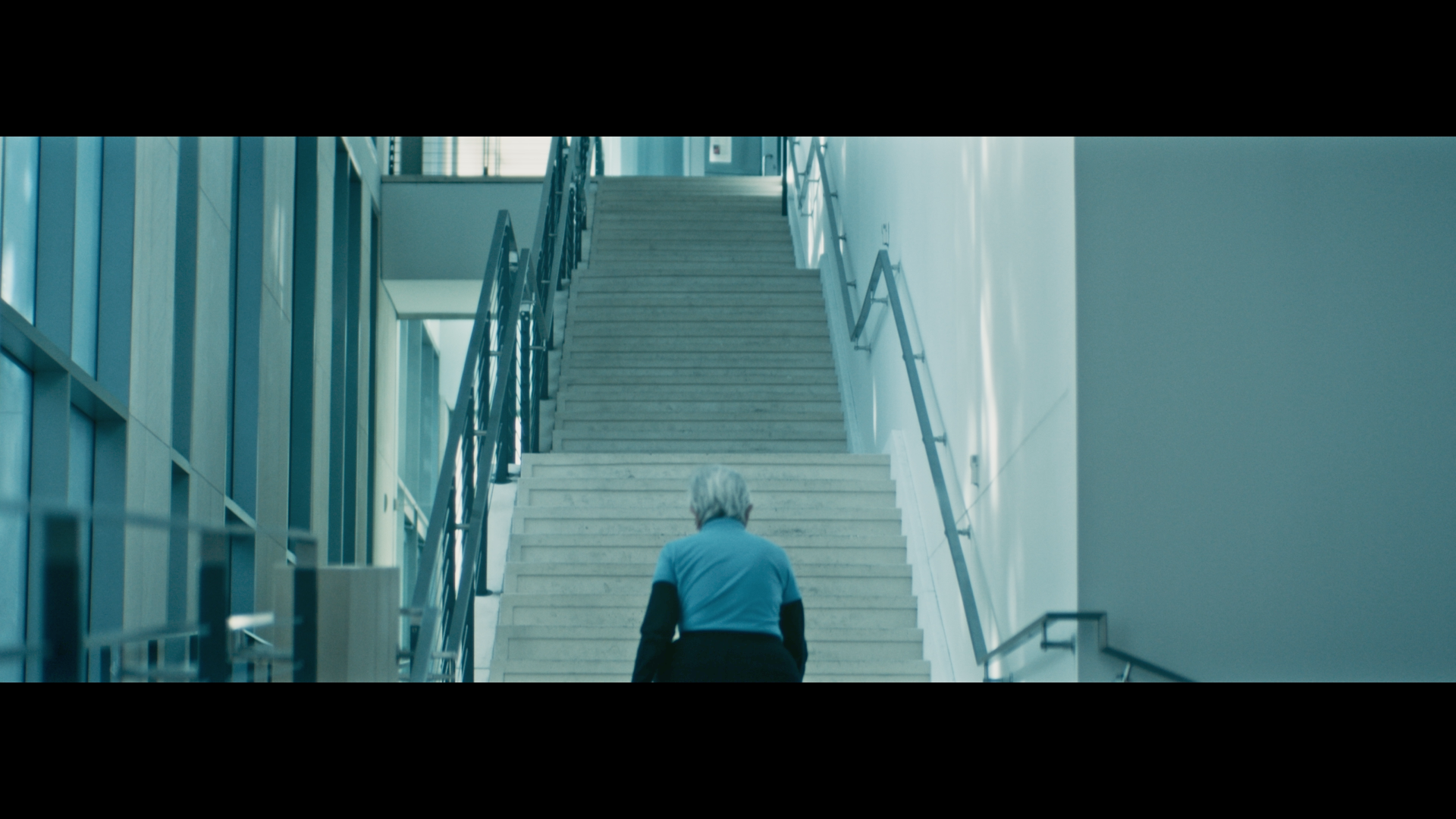Older person with gray hair walking up a large, modern staircase in a bright building with glass windows and metal railings.